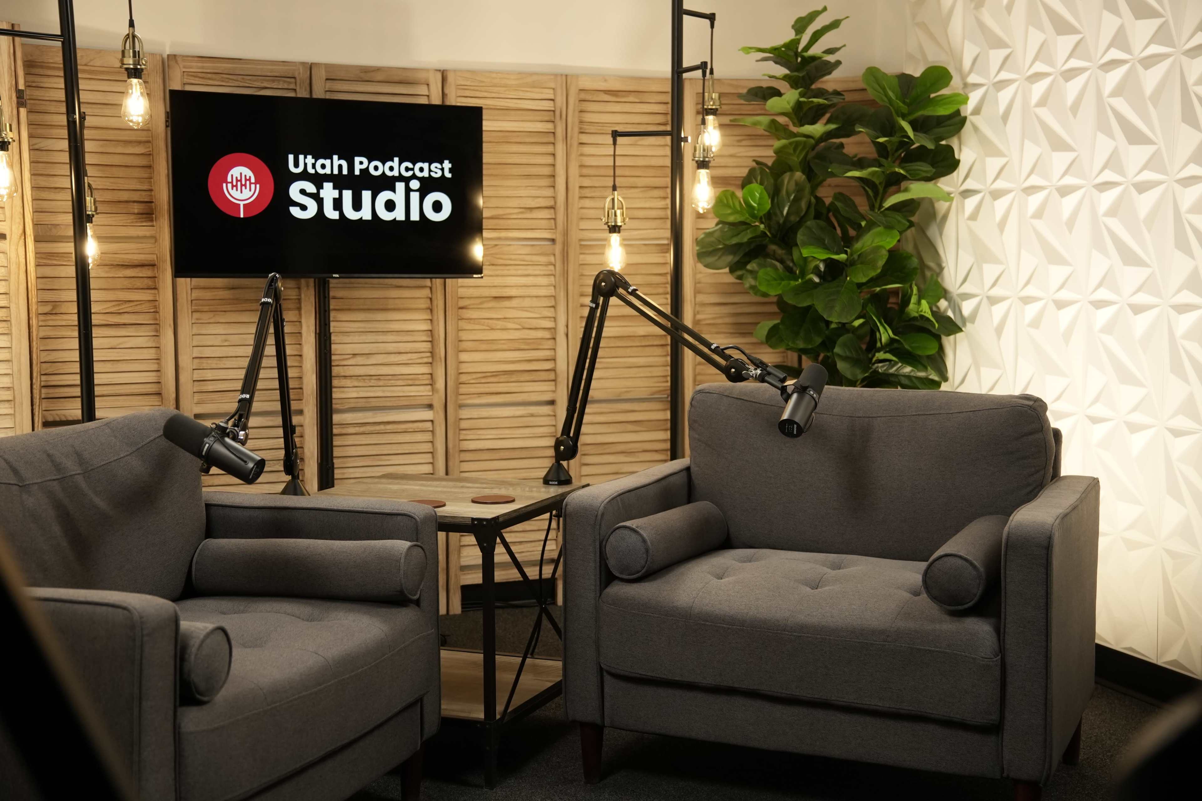 The image shows a podcast studio setup with two gray armchairs, a small wooden table, and a backdrop featuring the text "Utah Podcast Studio" illuminated by overhead lights.
