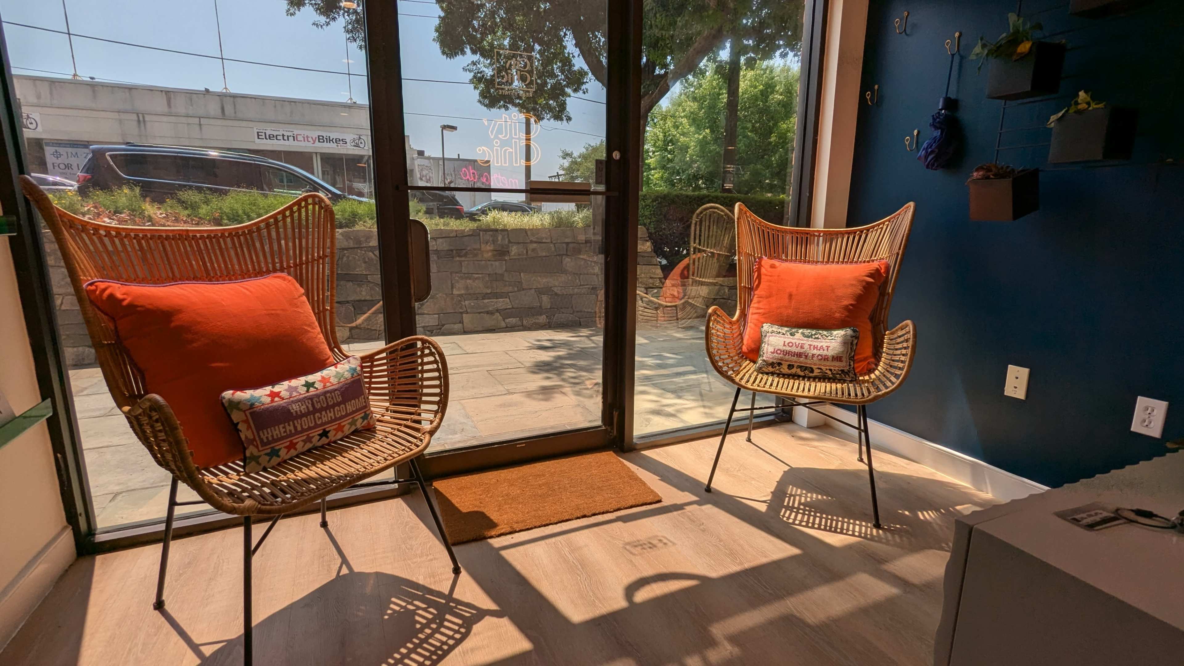 Two rattan chairs with orange cushions are positioned near a large glass door, allowing natural light to illuminate the space.