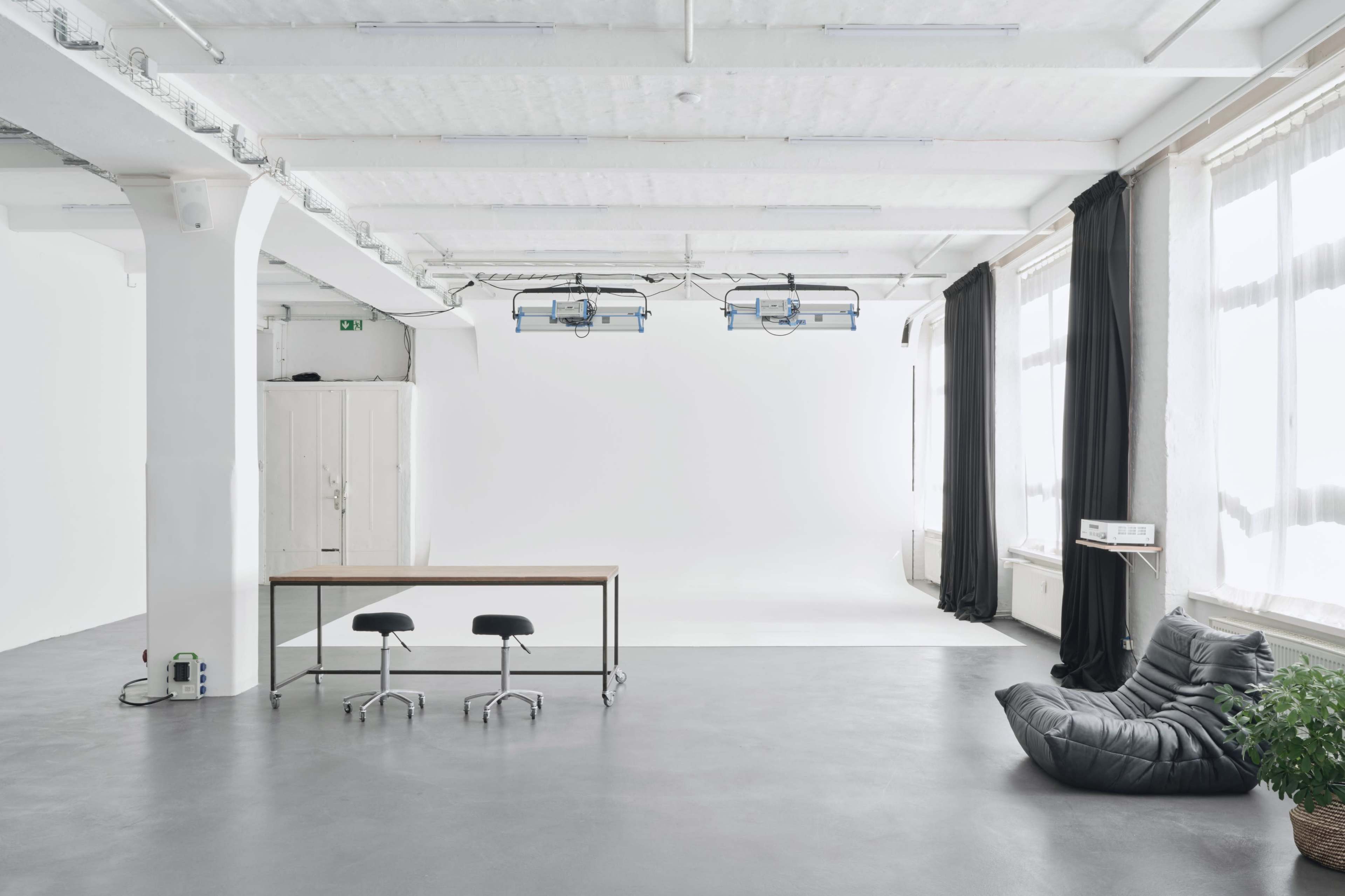 The image shows a spacious, well-lit studio with a large white wall, a wooden table with two chairs, and a cushioned seating area.