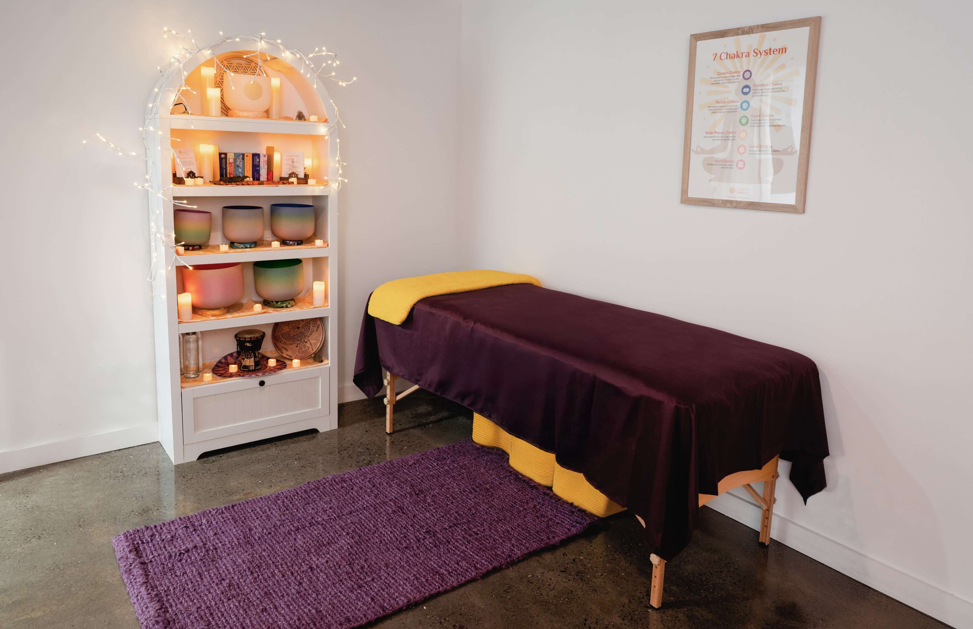 The image shows a calming therapy room featuring a massage table draped with a purple cover, a shelf with decorative bowls and ambient lighting, and a framed poster on the wall.