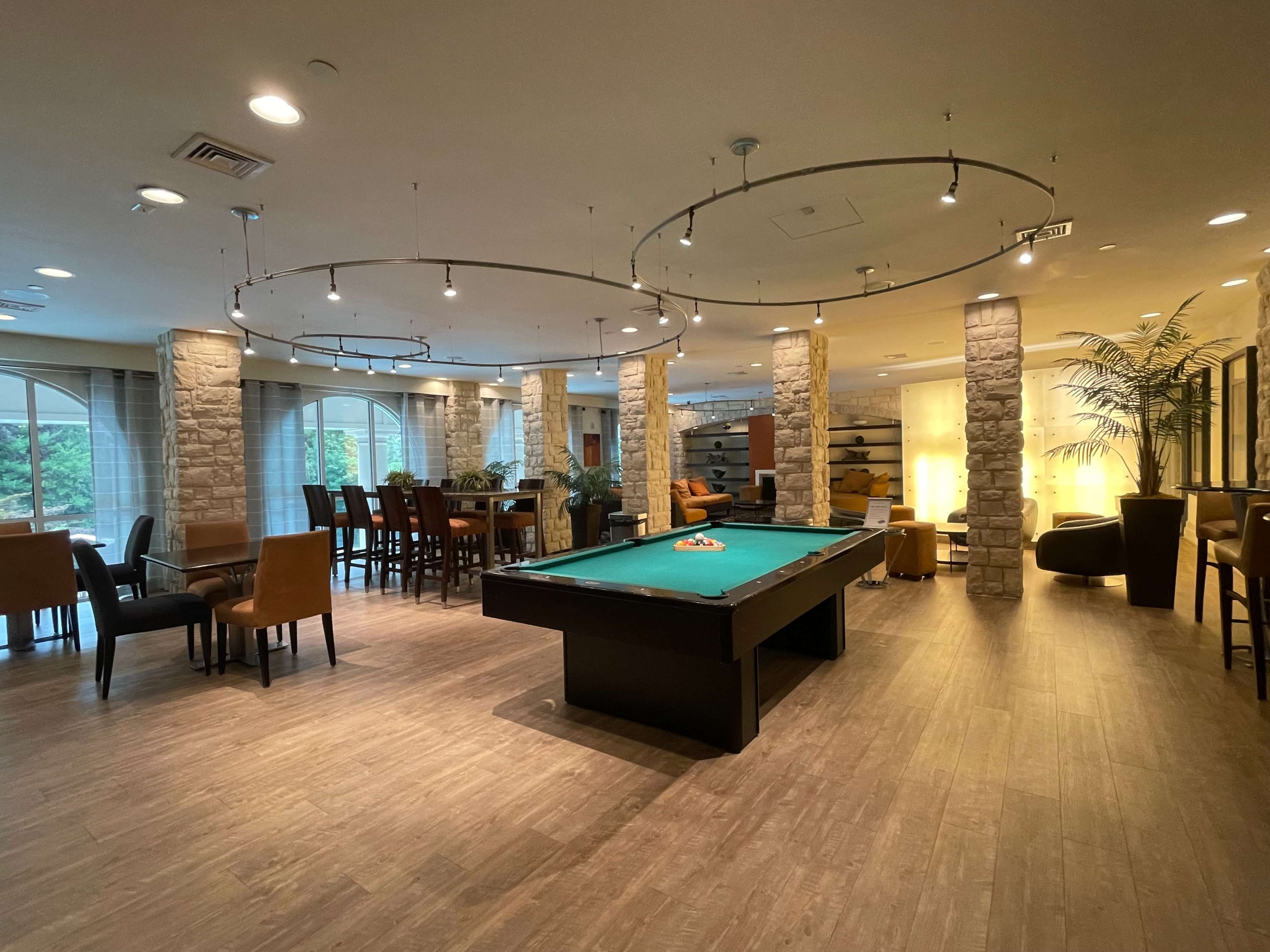 A spacious recreational lounge with a pool table at the center, surrounded by seating areas and illuminated by overhead lights.