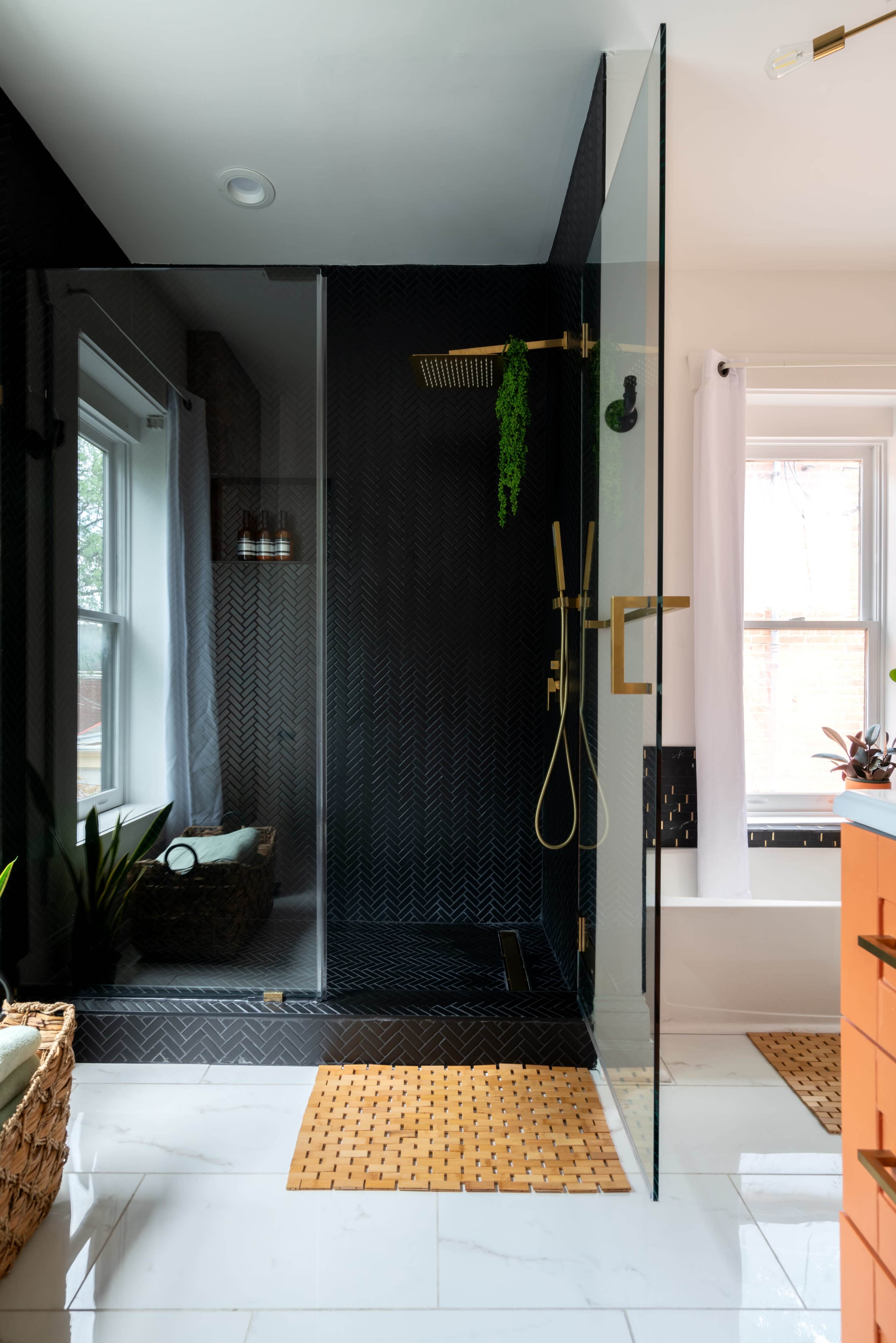 A modern shower area features a glass enclosure, a rainfall showerhead, and plants beside a window, with a woven mat on the marble floor.
