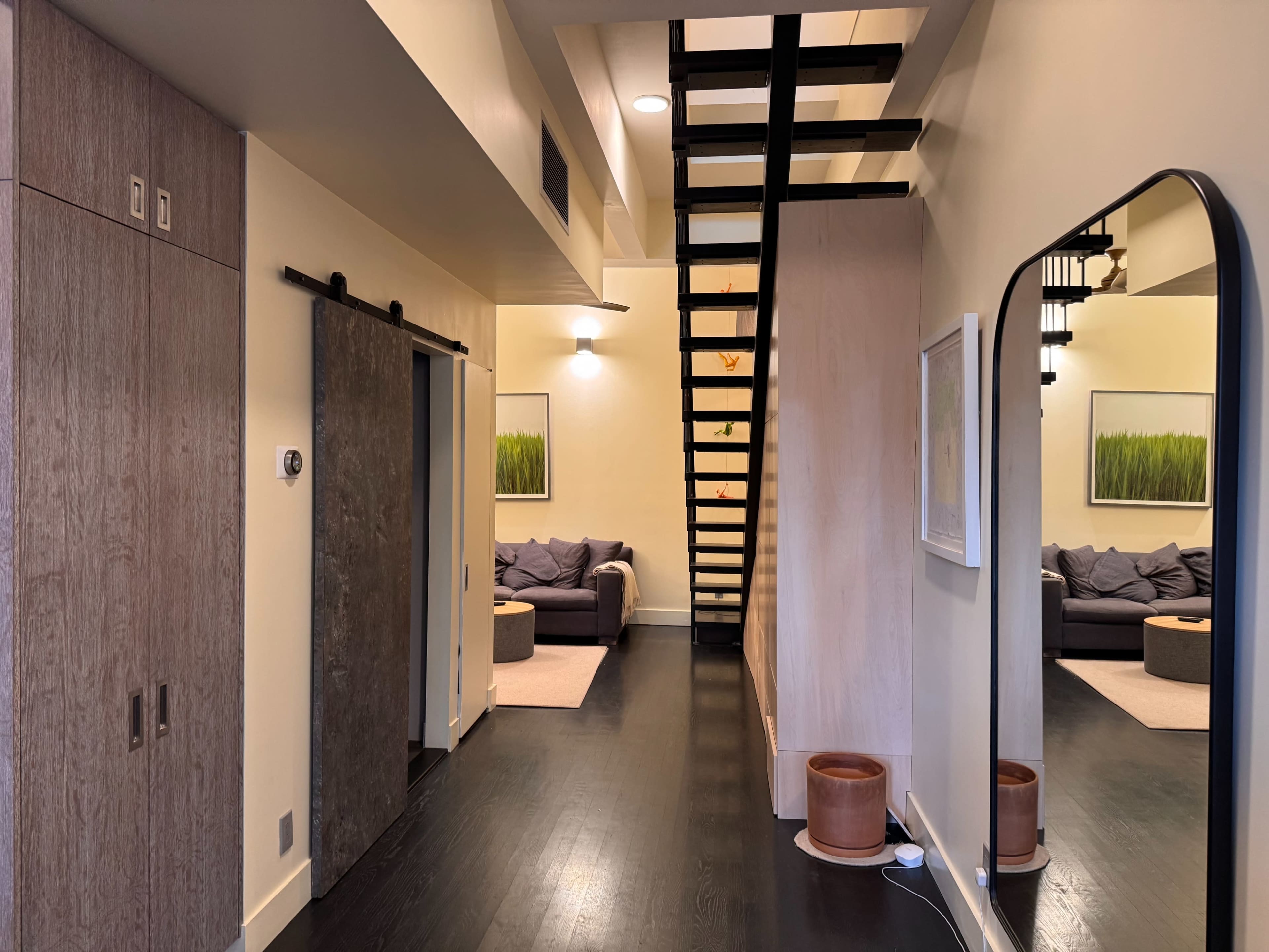 The image shows a modern interior featuring a staircase, a large mirror, and a cozy seating area with a sofa and decorative artwork on the walls.