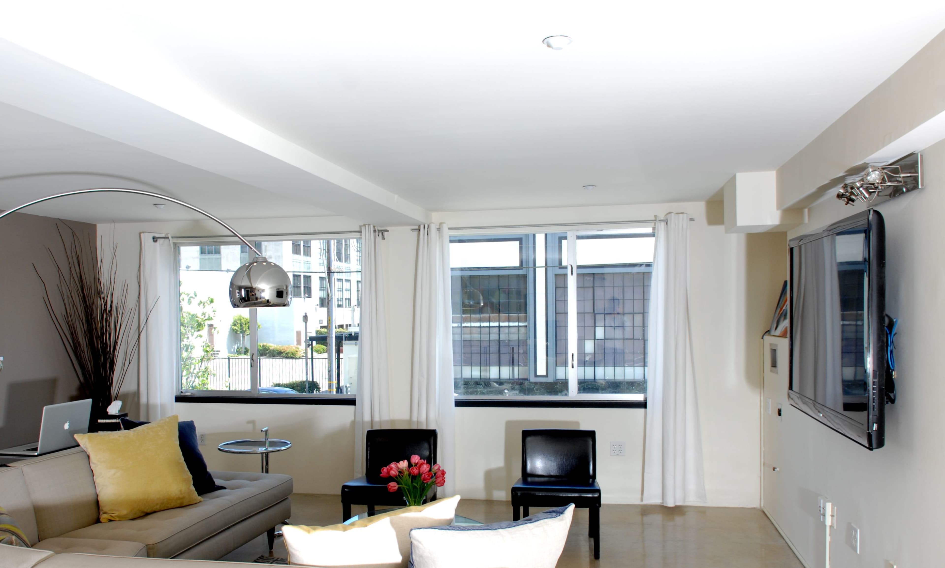 A modern living room with a beige sofa, two black chairs, a coffee table, and large windows allowing natural light.