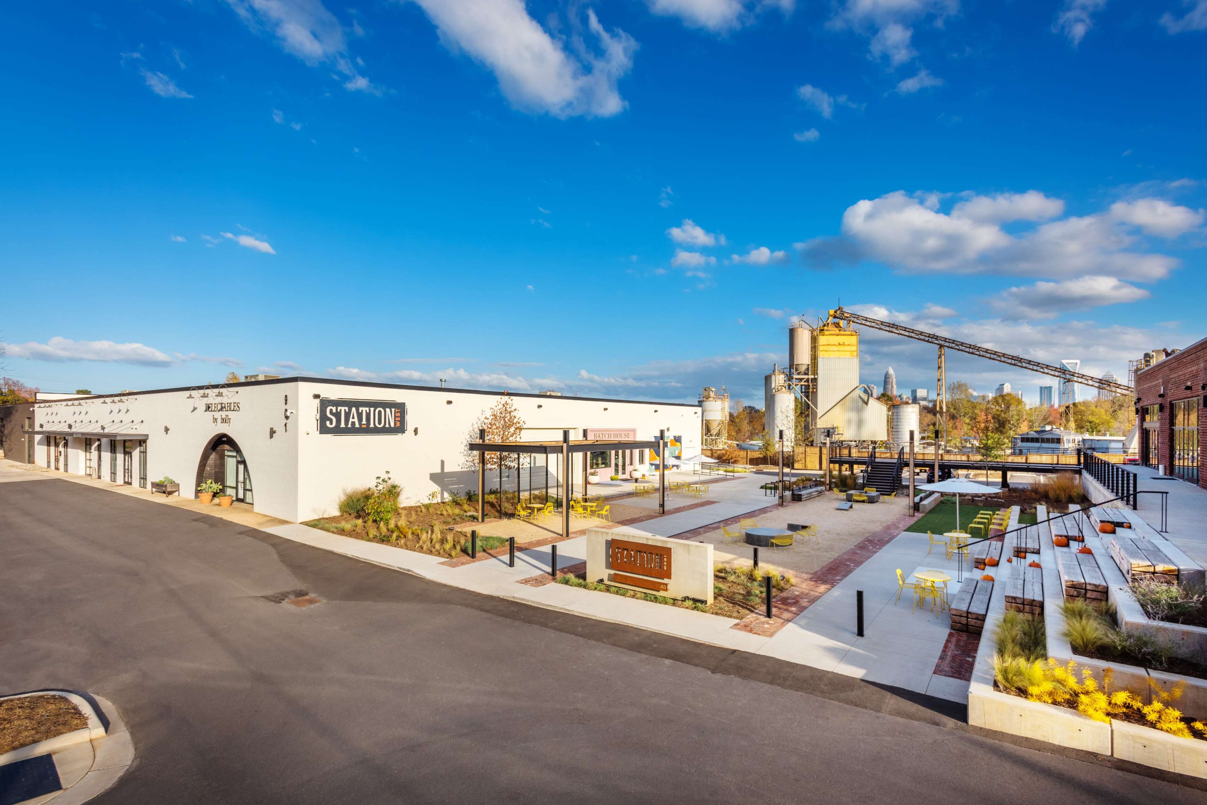 A modern commercial complex with white buildings, industrial silos, and landscaped outdoor seating areas under a blue sky.