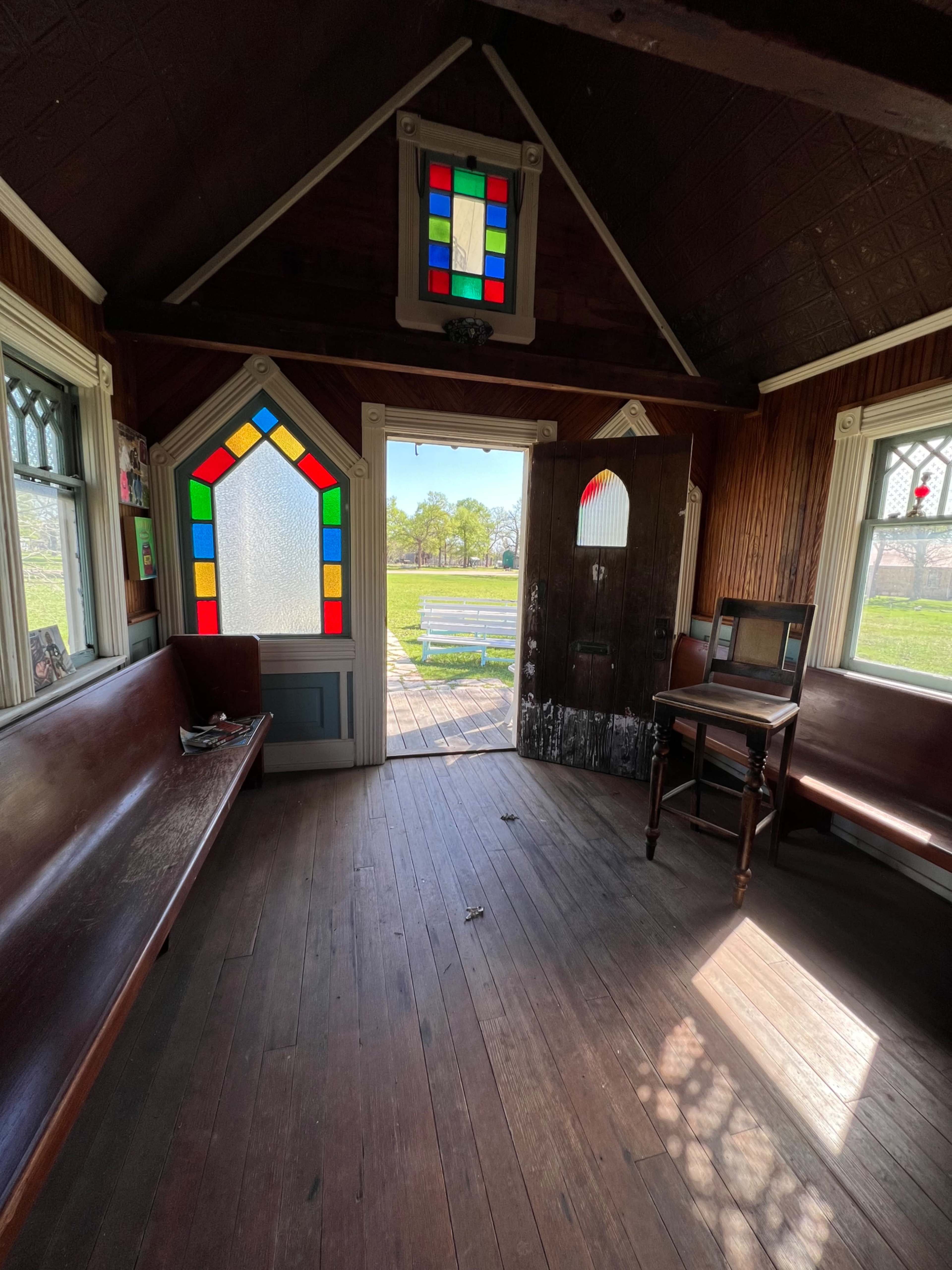 The interior of a small wooden structure features stained glass windows and simple furniture, with a view of a park visible through the open door.