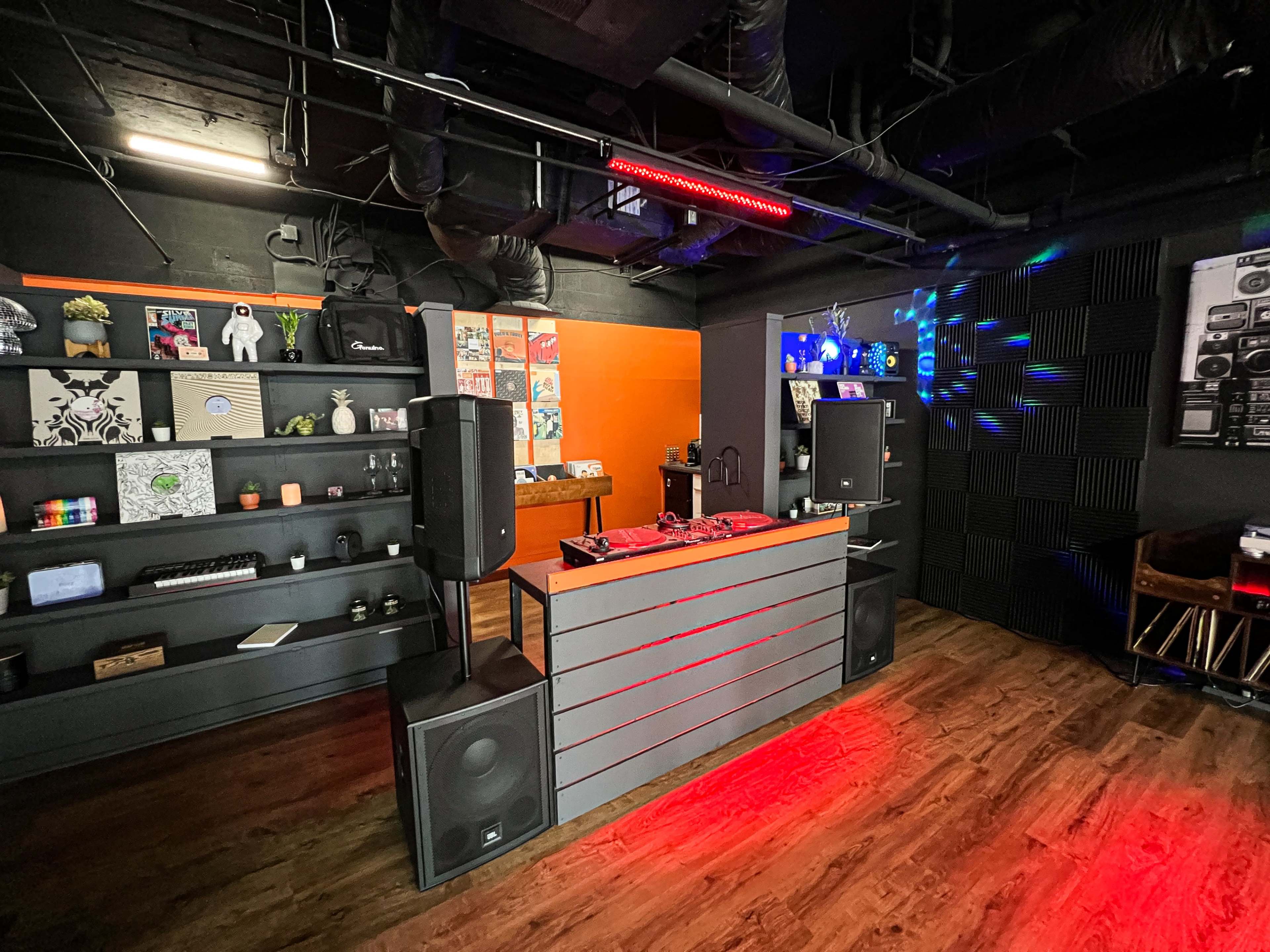 A modern music studio or recording space with a DJ setup, shelves displaying decor, and colorful lighting effects.