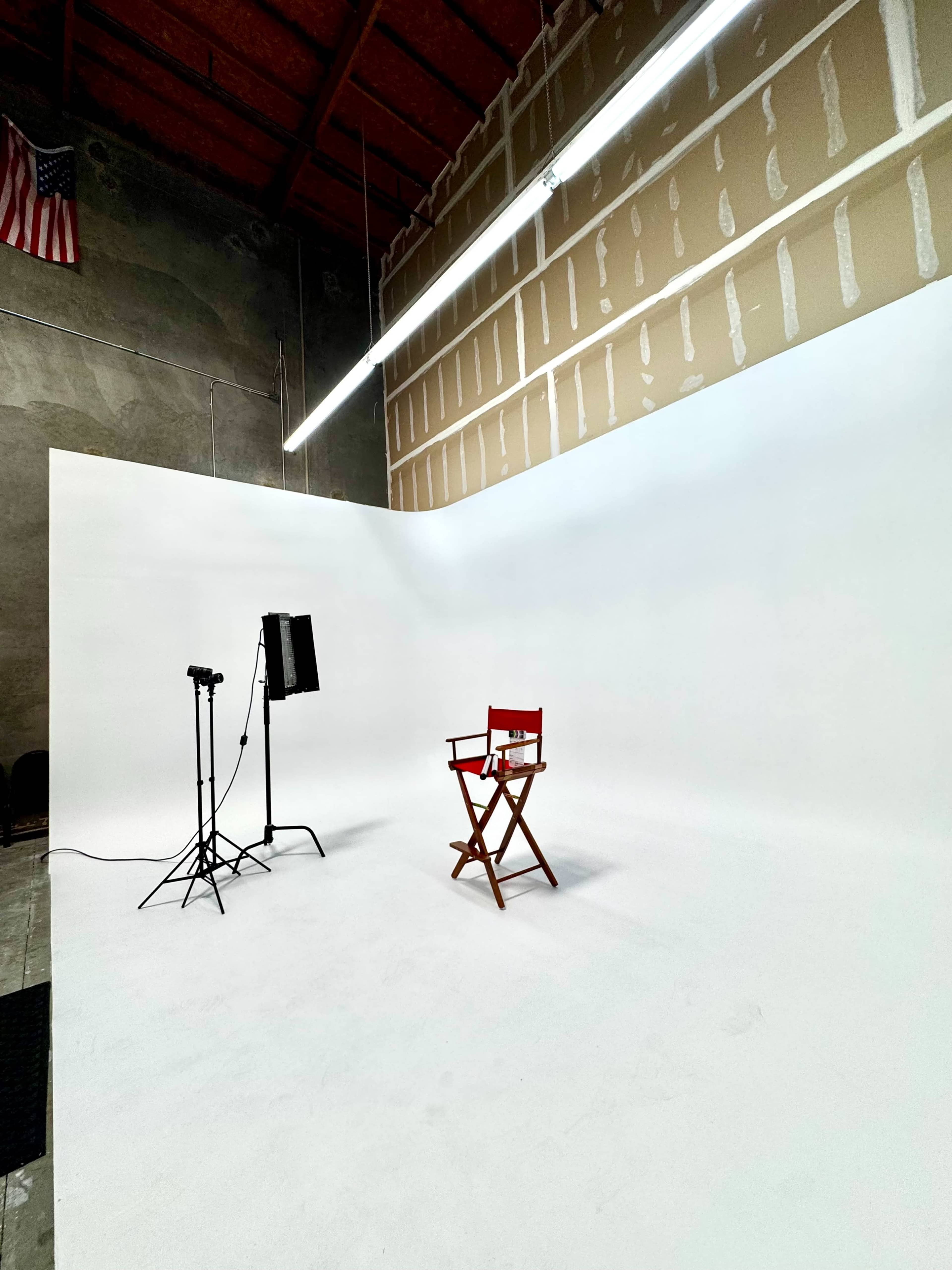 The image shows a minimalist studio with a white backdrop, a director's chair, and lighting equipment.