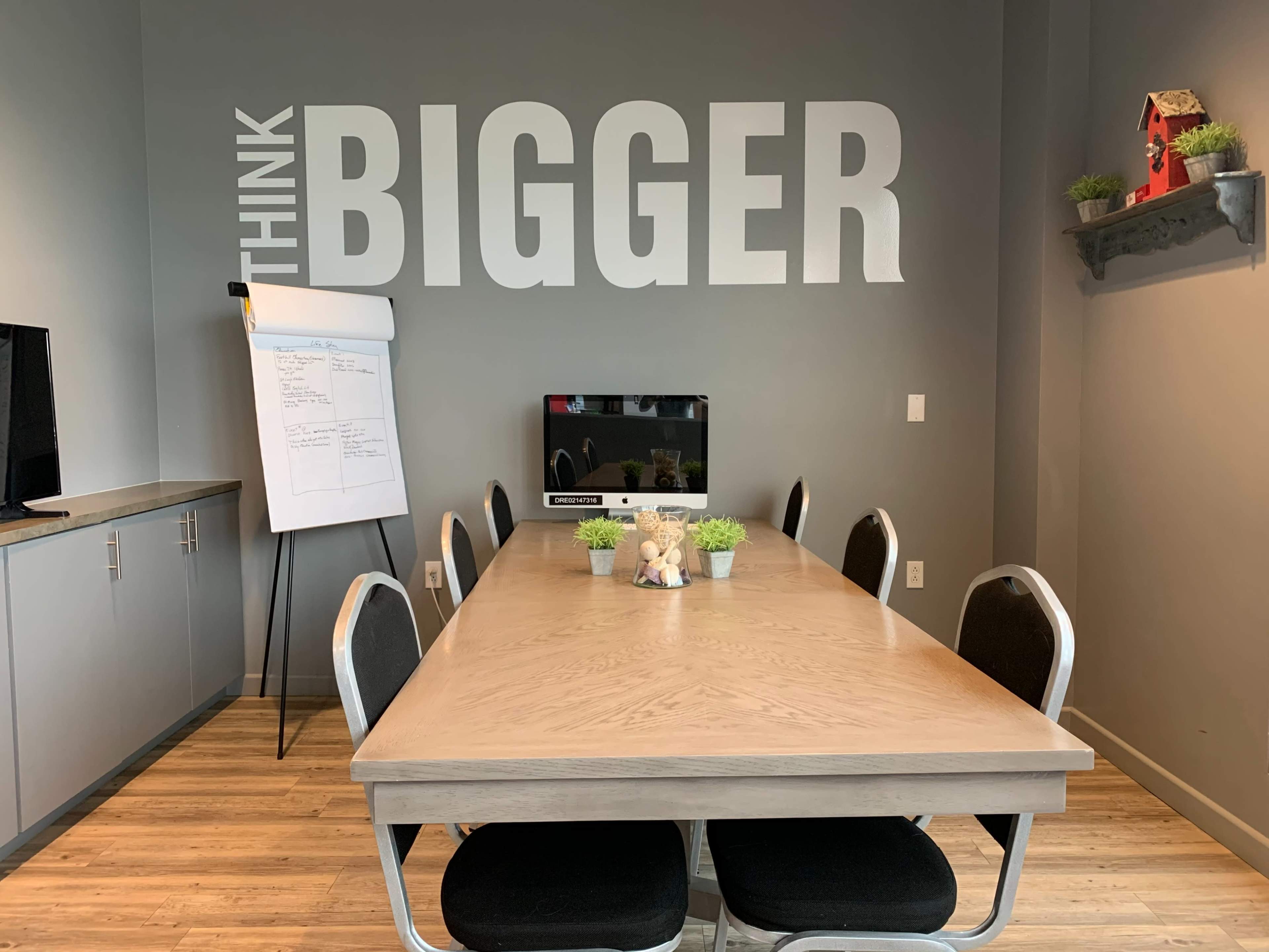 A conference room features a large wooden table surrounded by black chairs, a whiteboard on an easel, a television on the wall, and the phrase "THINK BIGGER" displayed prominently on the back wall.