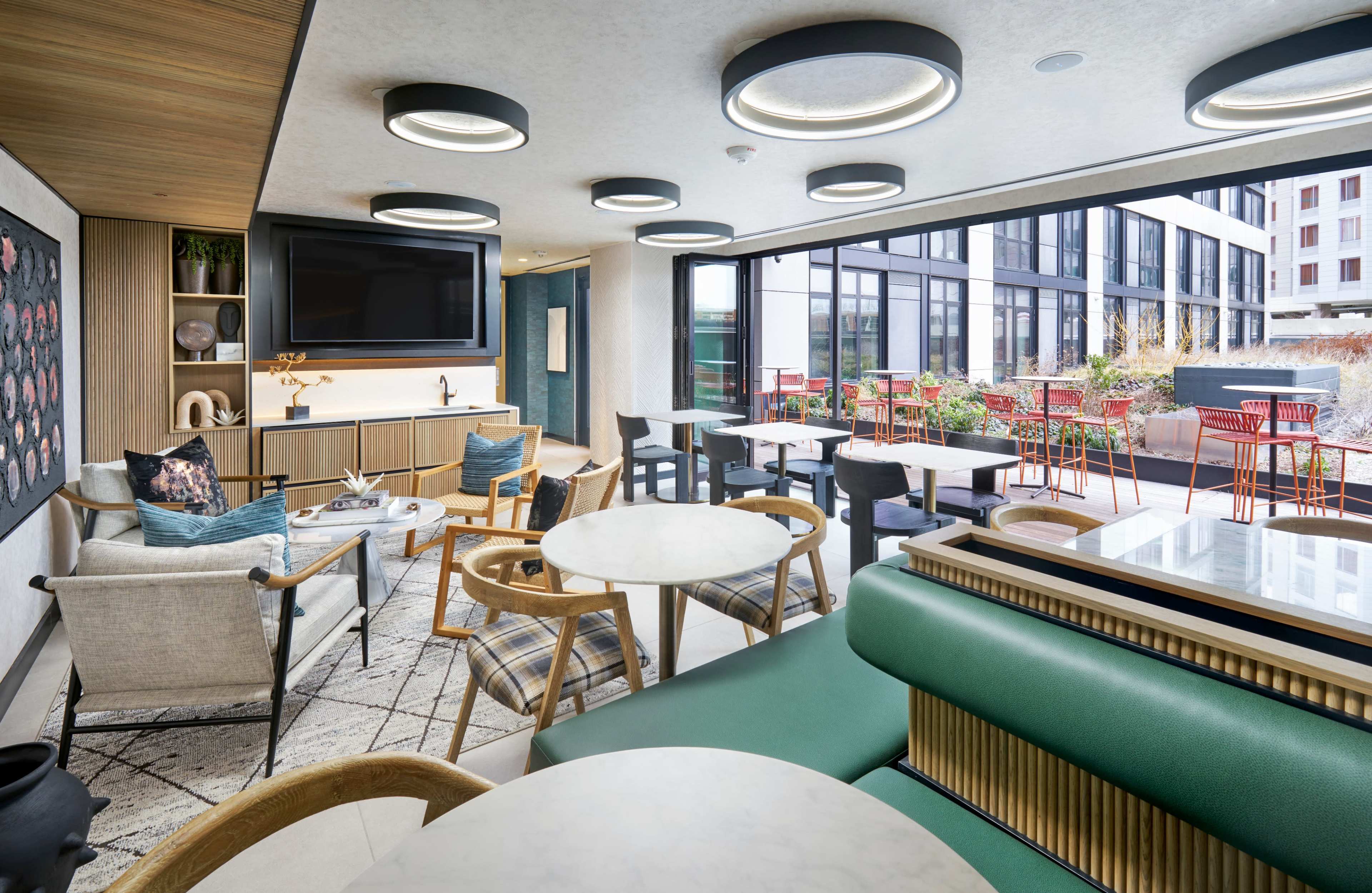 The image shows a modern lounge area with seating, a television, and large windows overlooking an outdoor patio with tables and chairs.