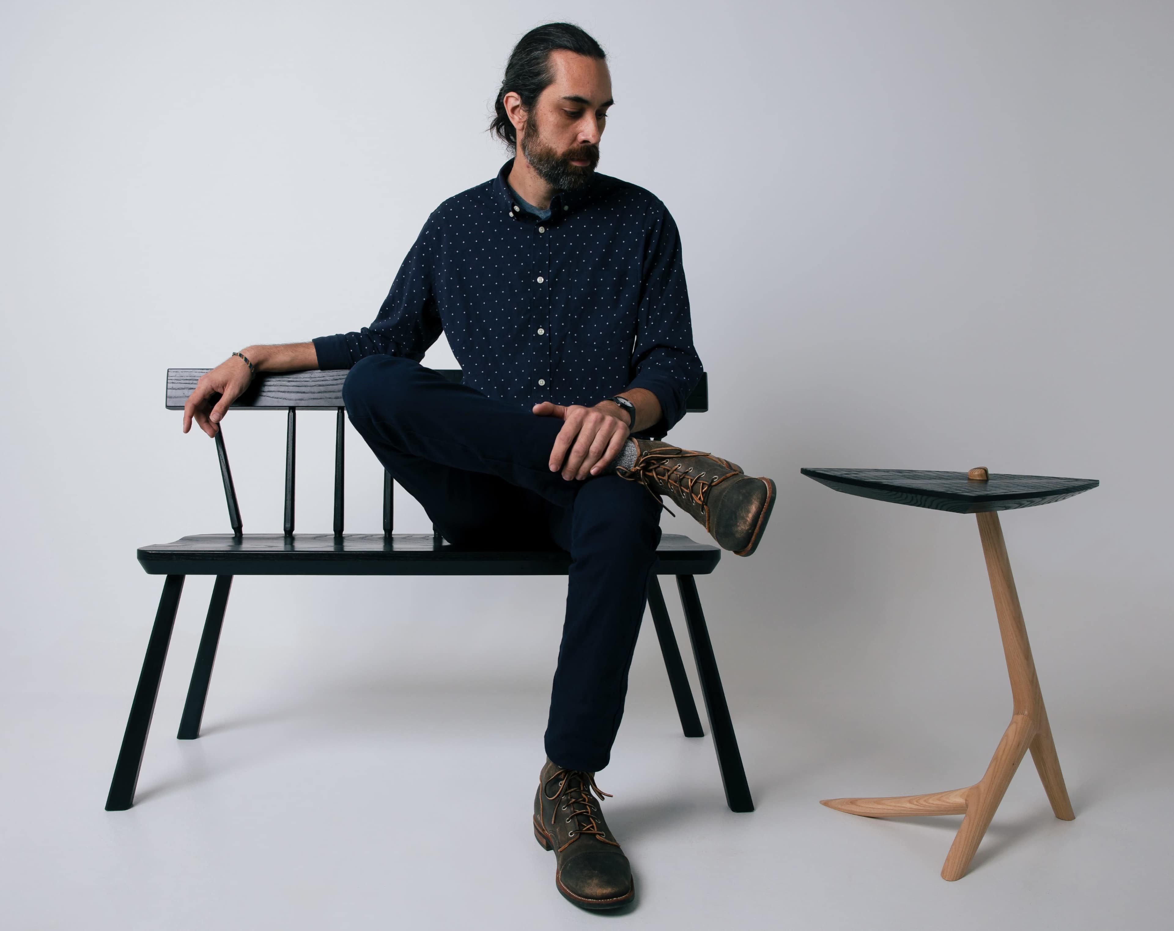 A man sits on a black bench with one leg crossed over the other, beside a small wooden side table.