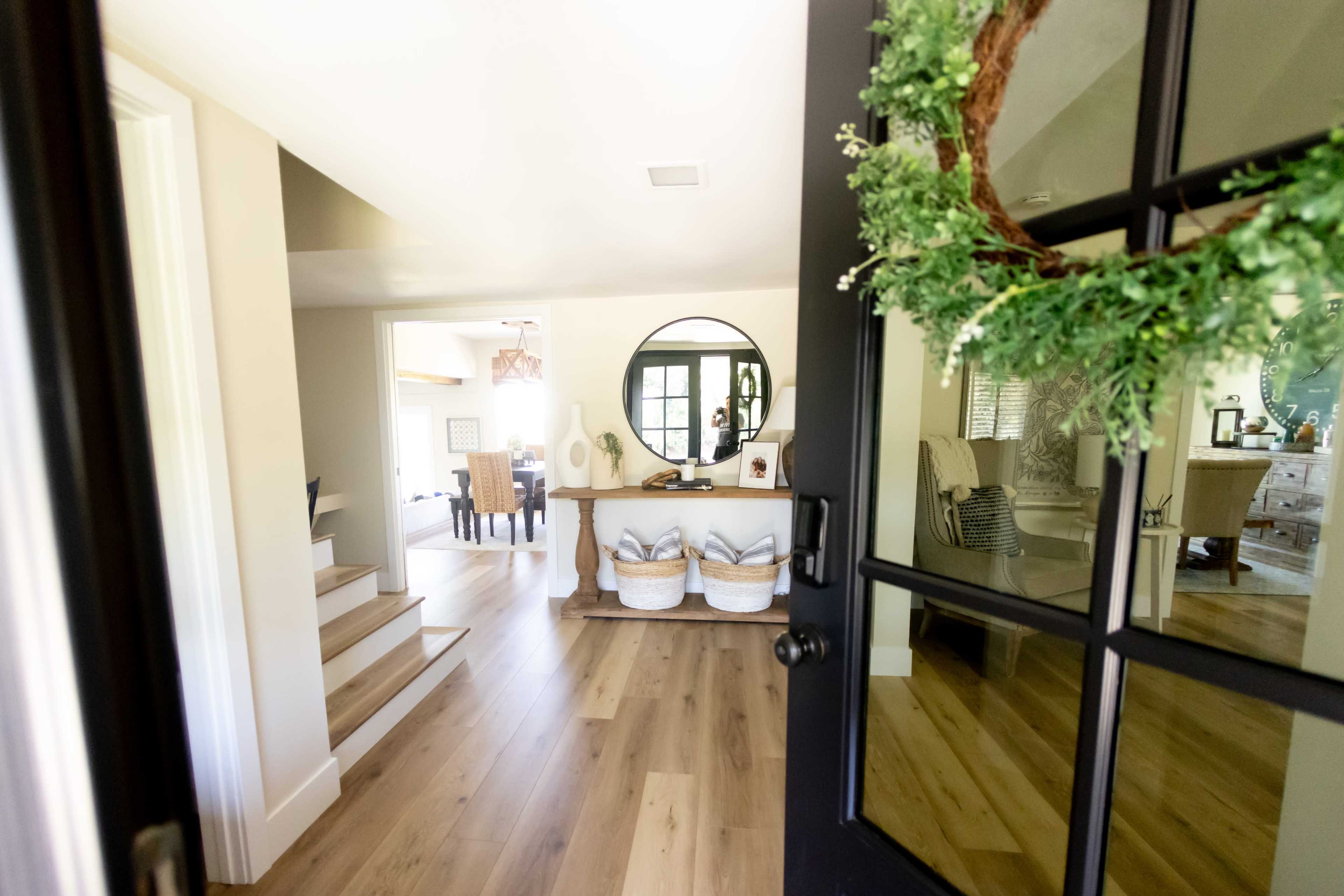 The image depicts a front entrance with a black door that opens to a light-filled living area featuring a round mirror and a staircase.