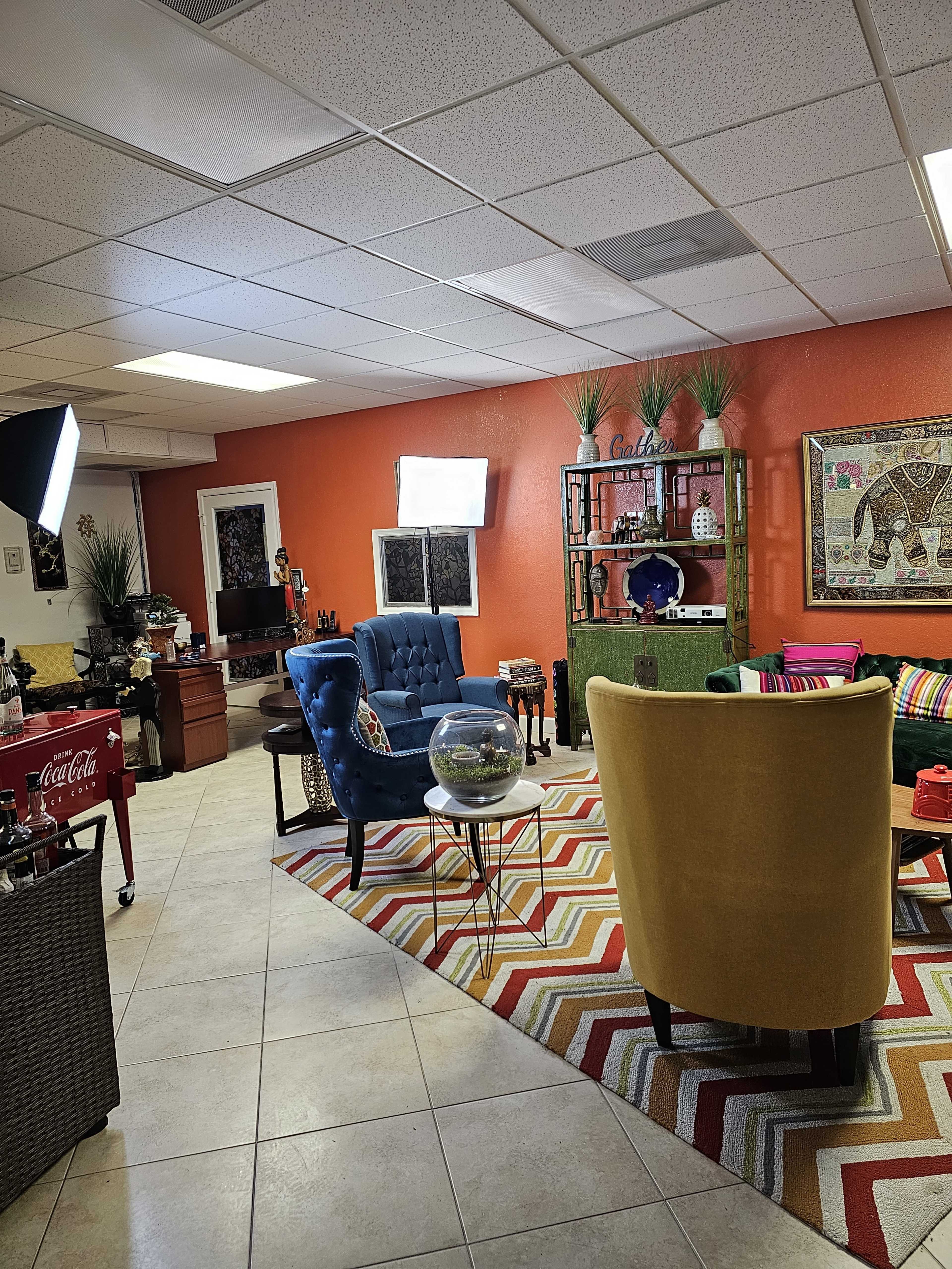 The image shows a vibrant, well-decorated lounge area with colorful furniture, a patterned rug, and shelves displaying decorative items.