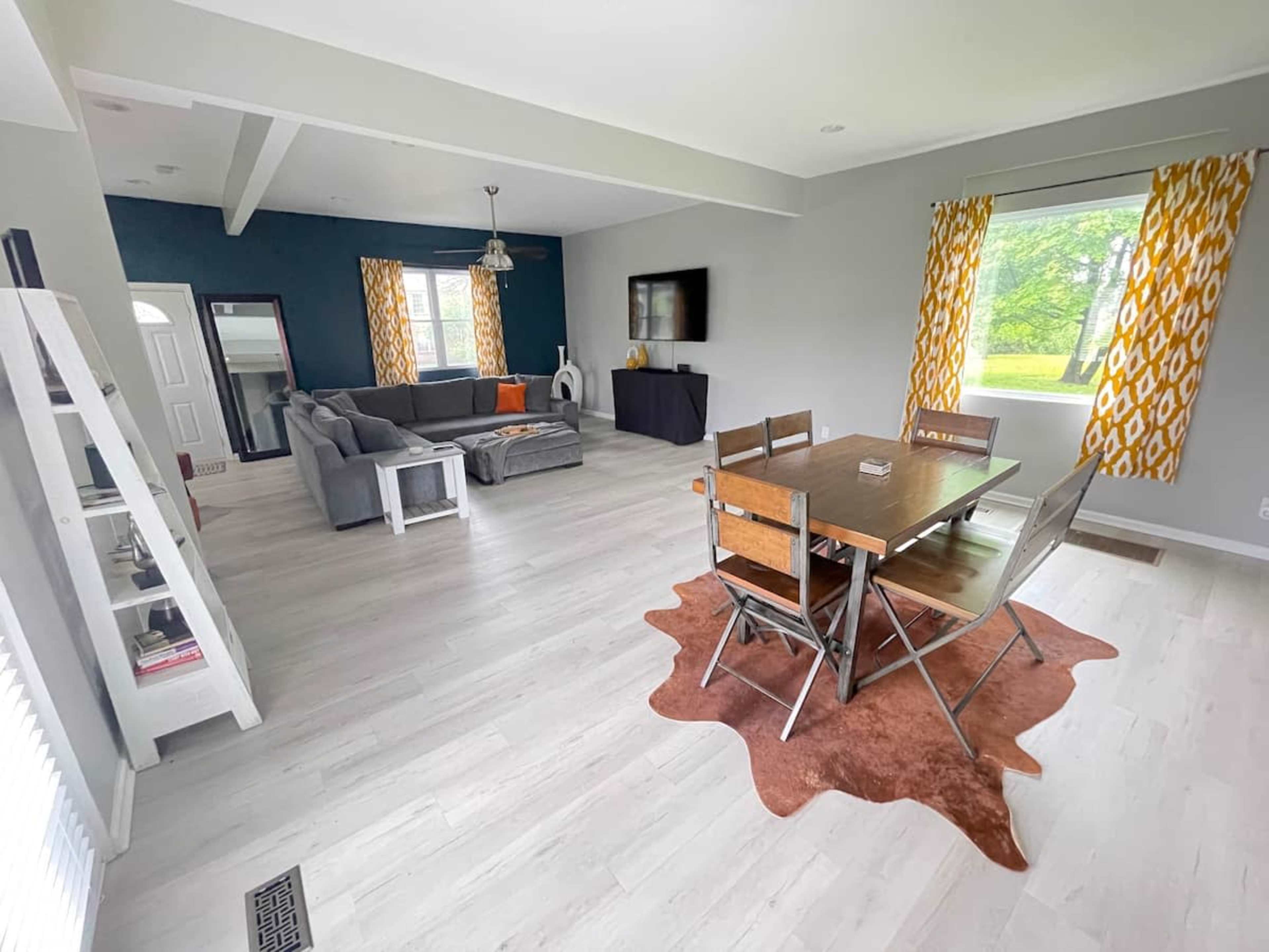 A modern, open-concept living and dining area featuring a gray sofa, a wooden dining table with metal chairs, and large windows with patterned yellow curtains.