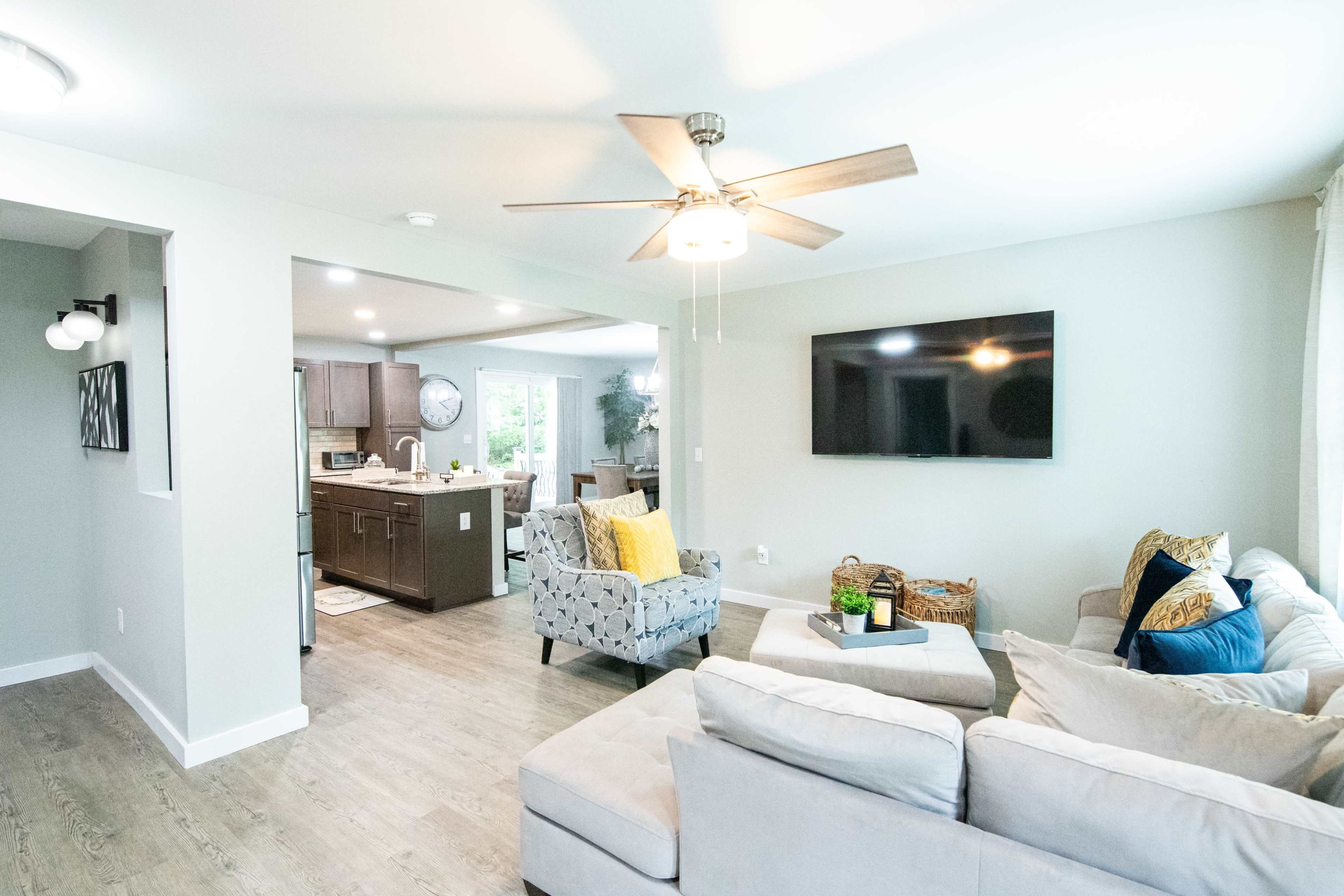A modern living room with a ceiling fan, a large television mounted on the wall, and seating arrangements that include a light gray sectional sofa and patterned accent chairs, with a view of an adjacent kitchen.
