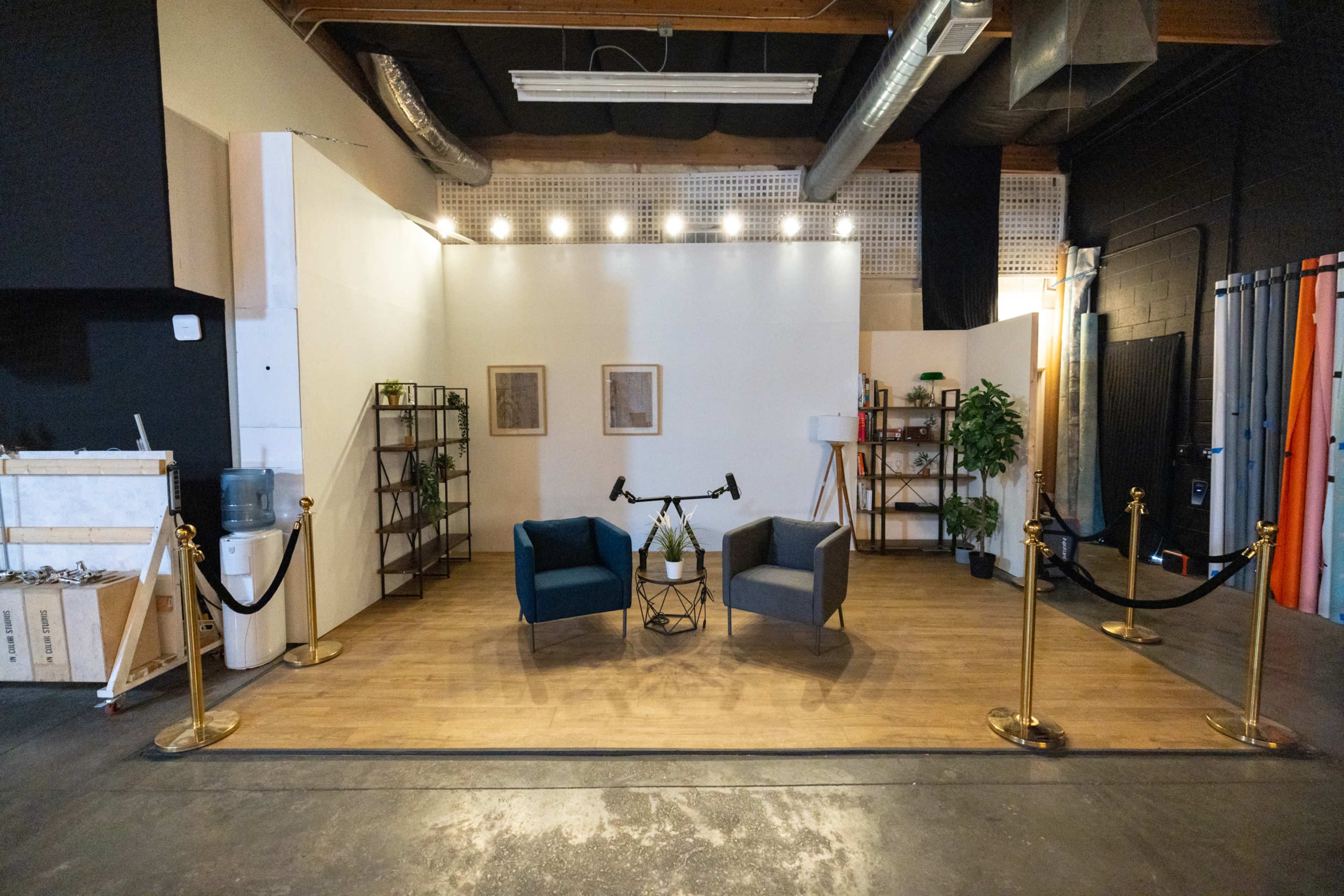 The image shows a minimalist studio setup featuring two chairs, a small table, and decorative plants, all arranged on a wooden floor with a backdrop of white walls and ambient lighting.