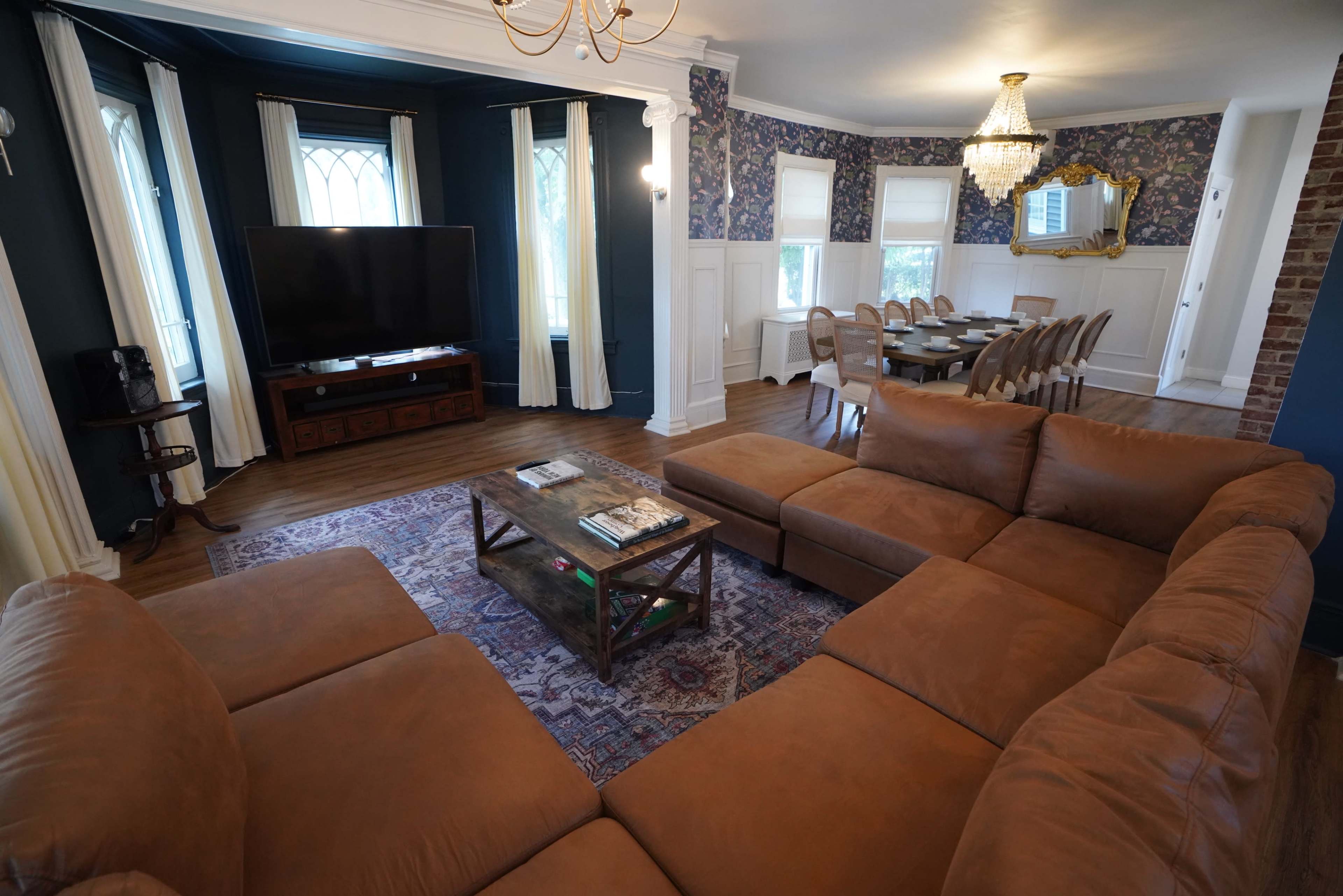 A spacious living room features a brown sectional sofa, a coffee table, a large television, and a dining area with a long table surrounded by chairs.