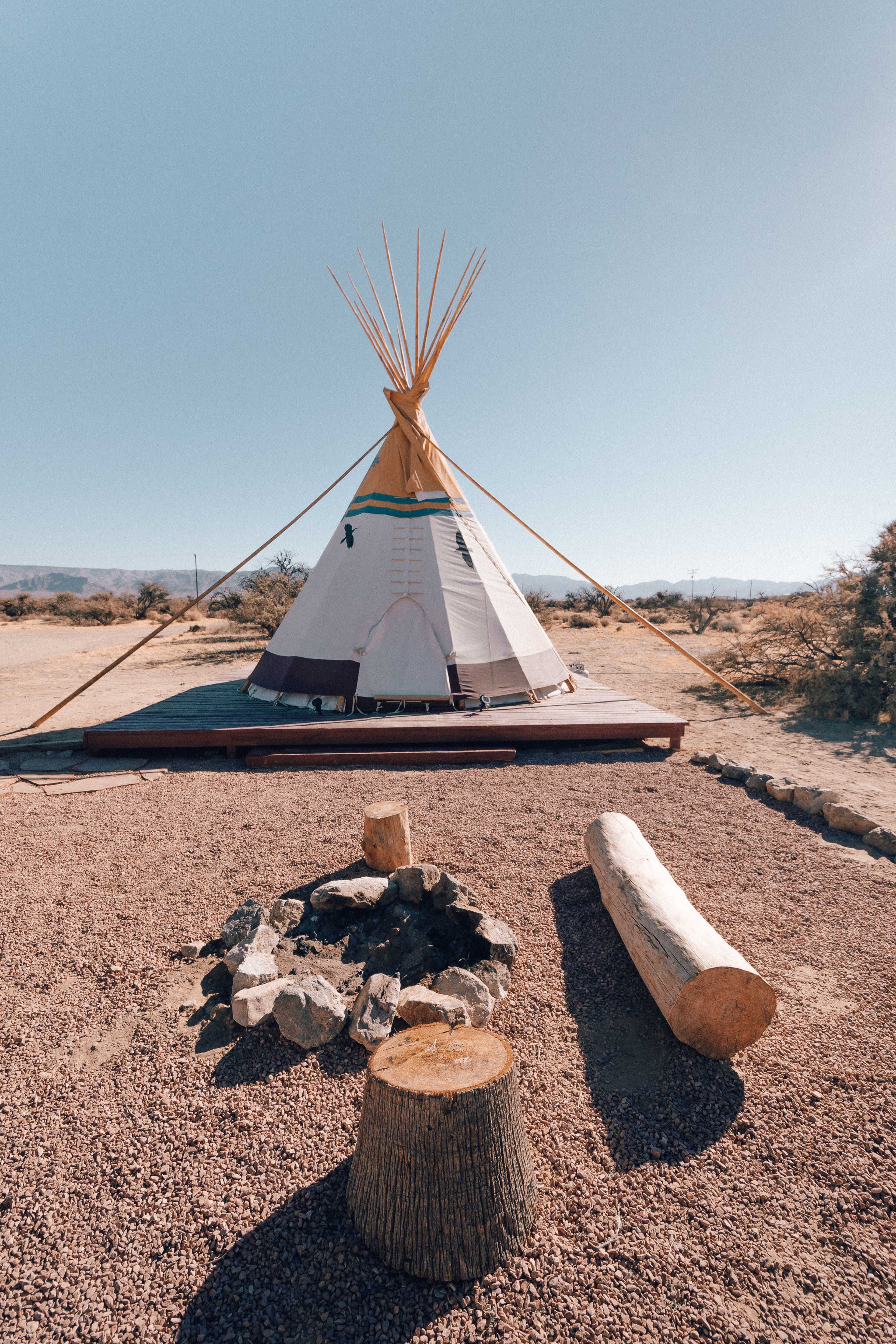 A teepee stands on a wooden platform in a desert landscape, surrounded by a circle of stones and a log near a fire pit.