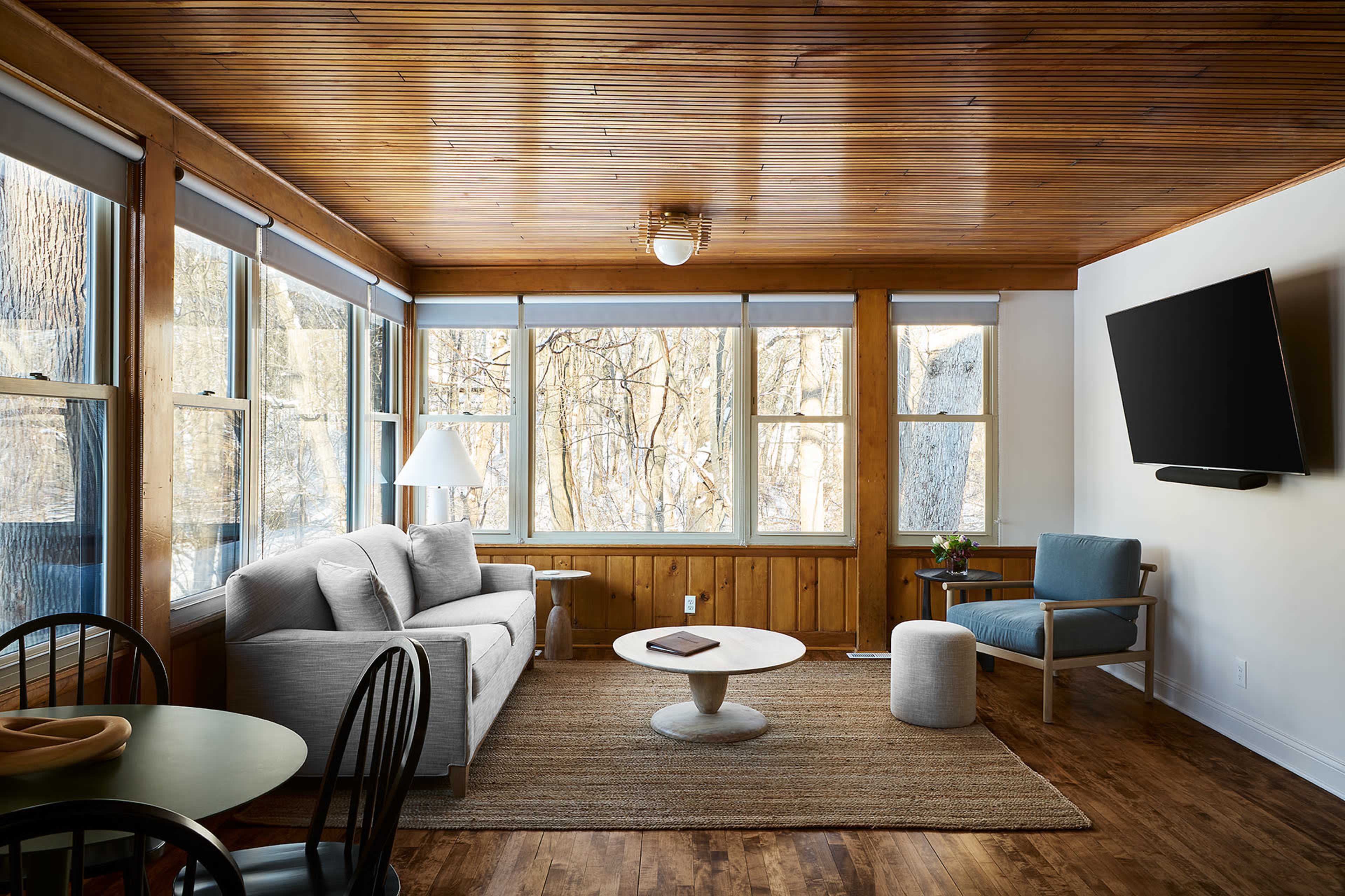 A sunlit living room featuring a gray sofa, light blue accent chair, round coffee table, and large windows overlooking trees.