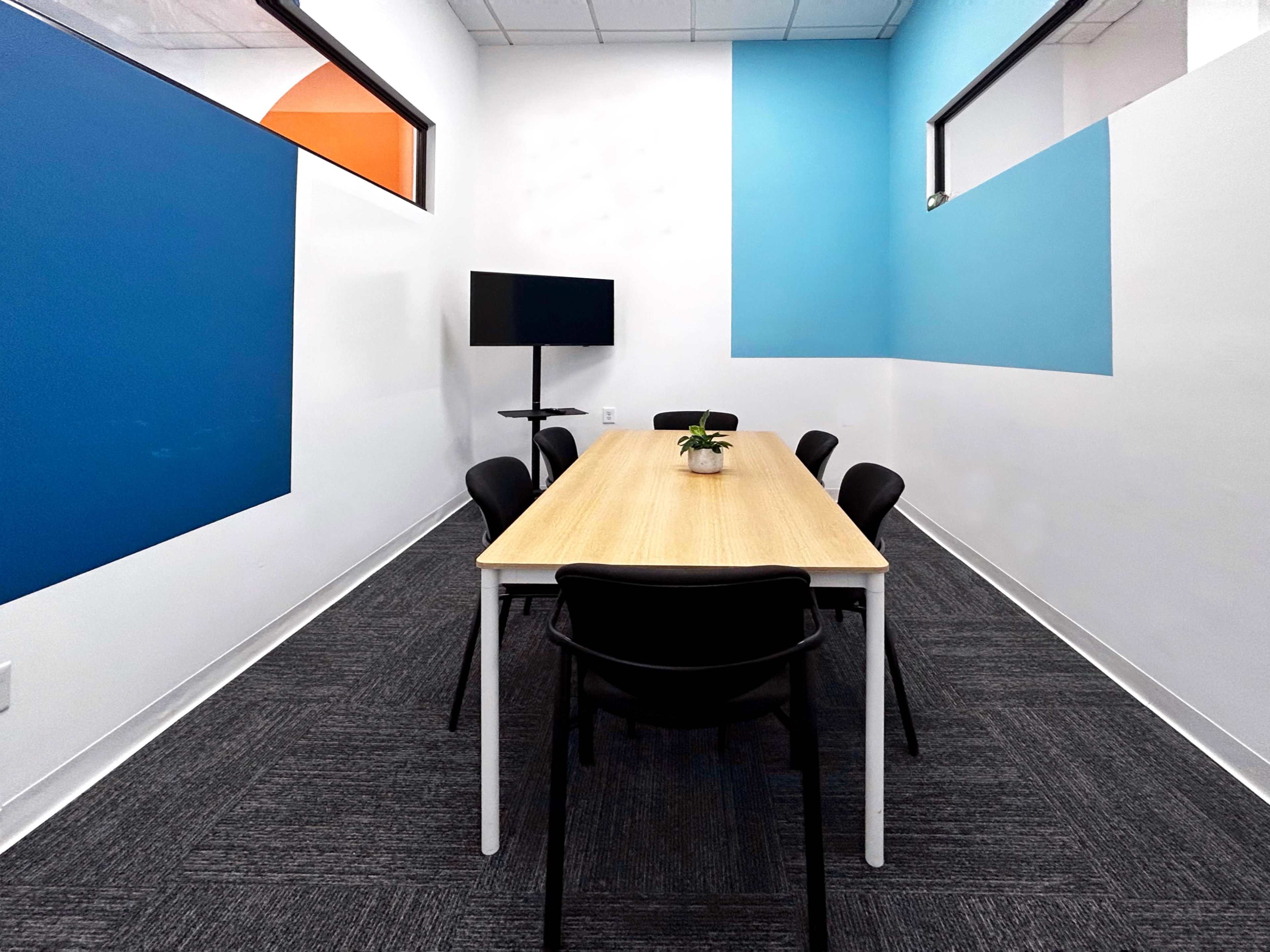 A conference room features a long wooden table surrounded by black chairs, with blue and orange accent walls and a television on a stand.