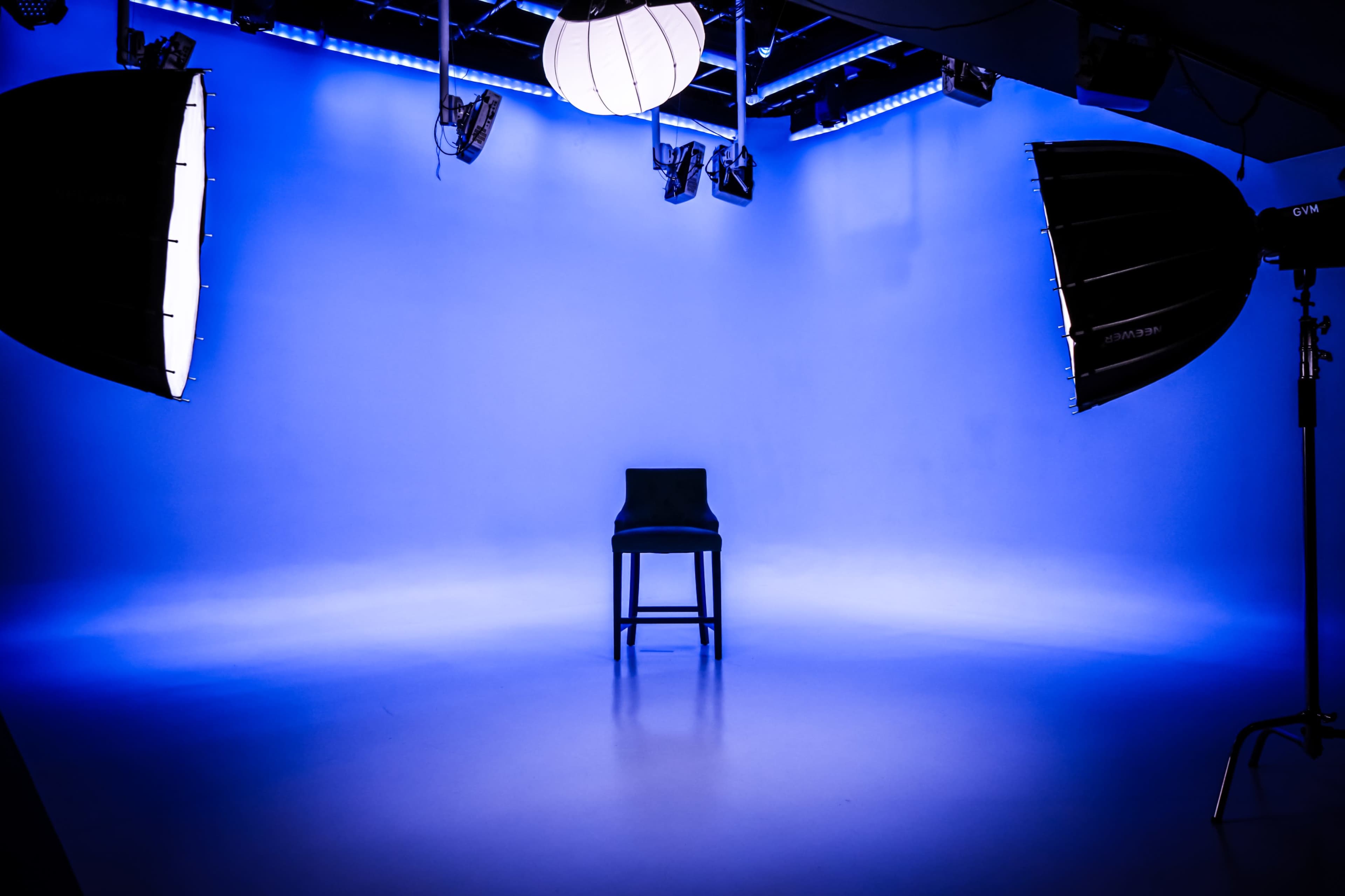 The image shows a minimalist studio setup featuring a single chair in the center, illuminated by two large softbox lights and a hanging lamp against a blue backdrop.