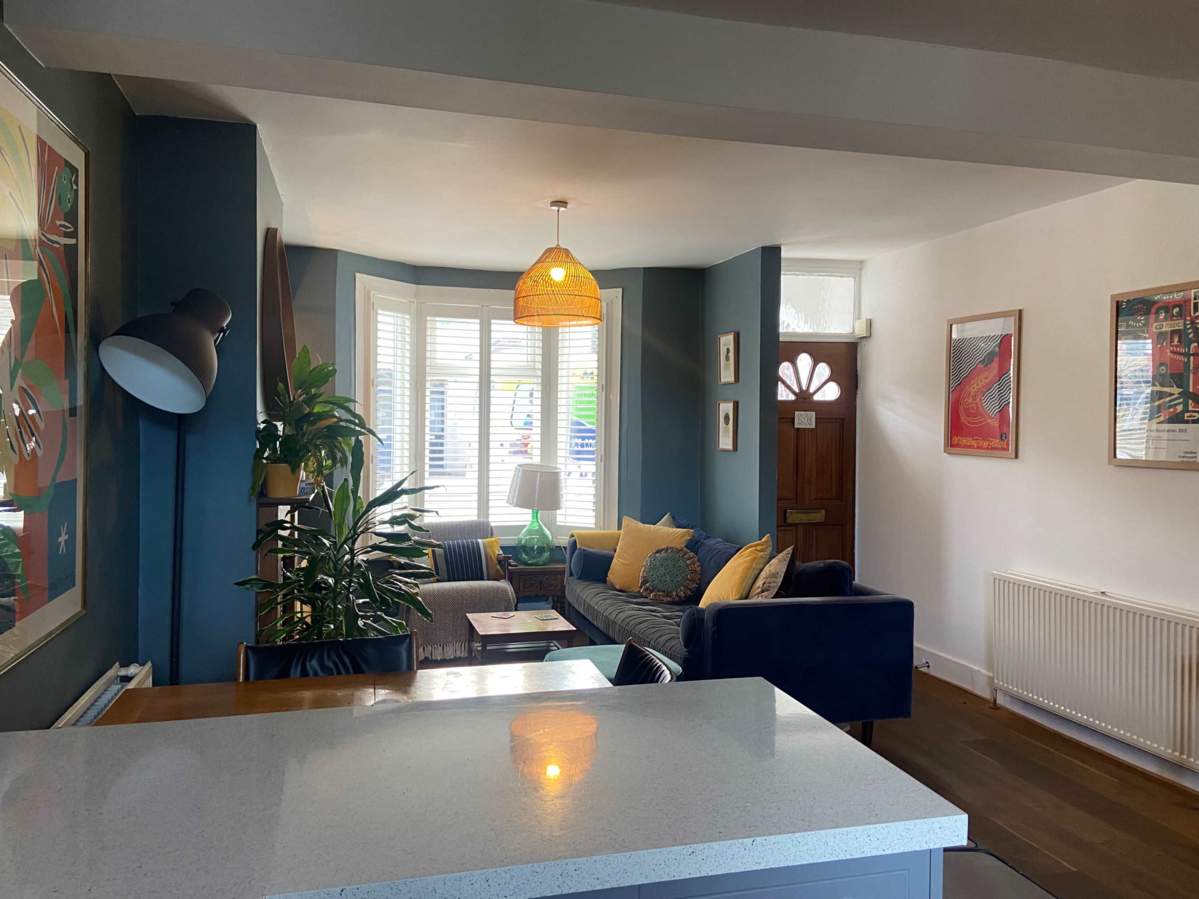 A cozy living room features a blue sofa, a plant, and artwork on the walls, with a door leading outside visible in the background.