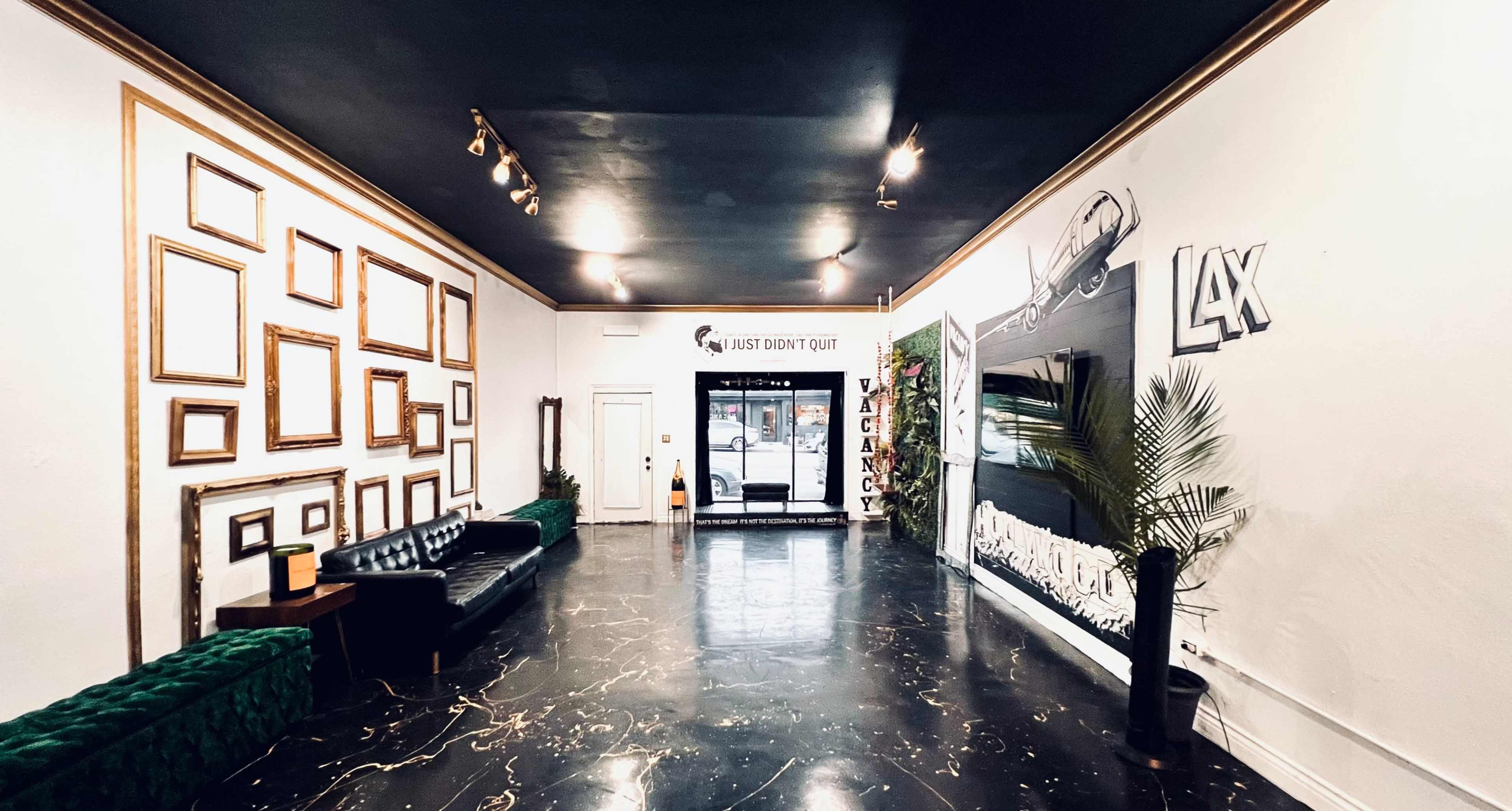 A spacious lobby with a black floor, framed artworks on the walls, a green velvet bench, a black leather sofa, and a doorway leading outside.