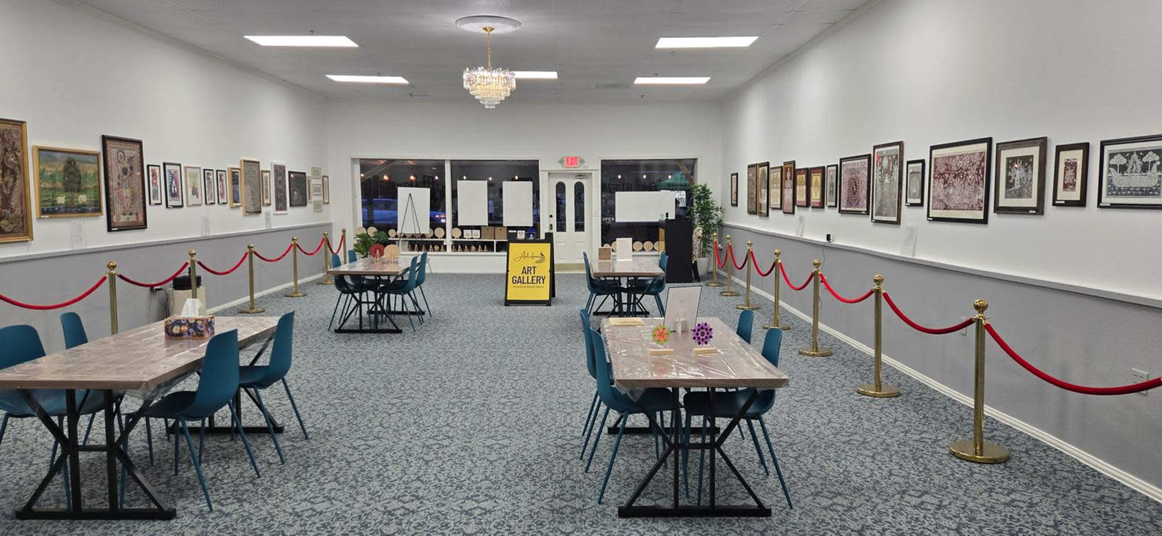 The image shows an empty gallery space with tables, chairs, and artwork displayed on the walls.
