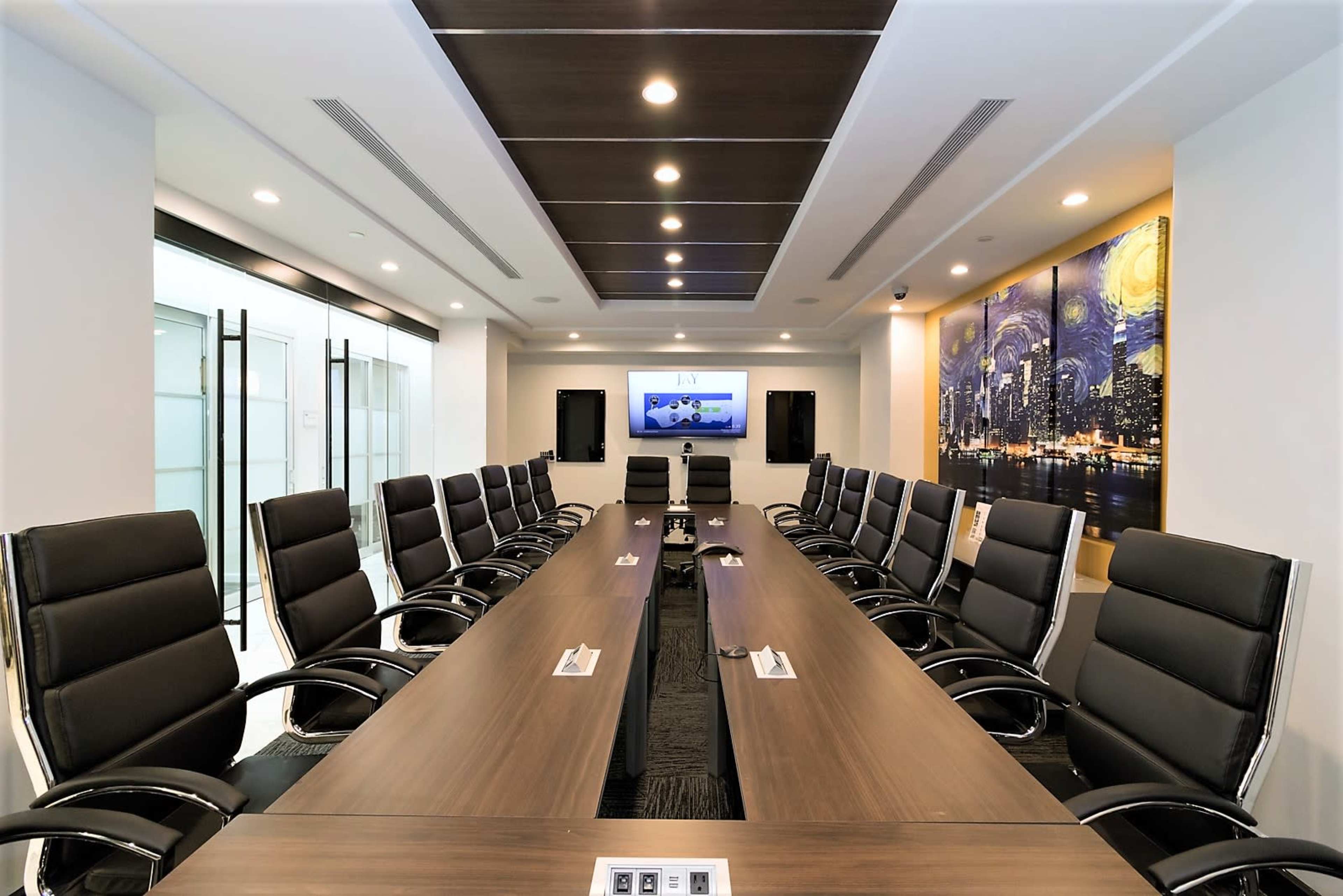 A modern conference room features a long wooden table surrounded by black leather chairs, with a large screen and a cityscape painting on the wall.