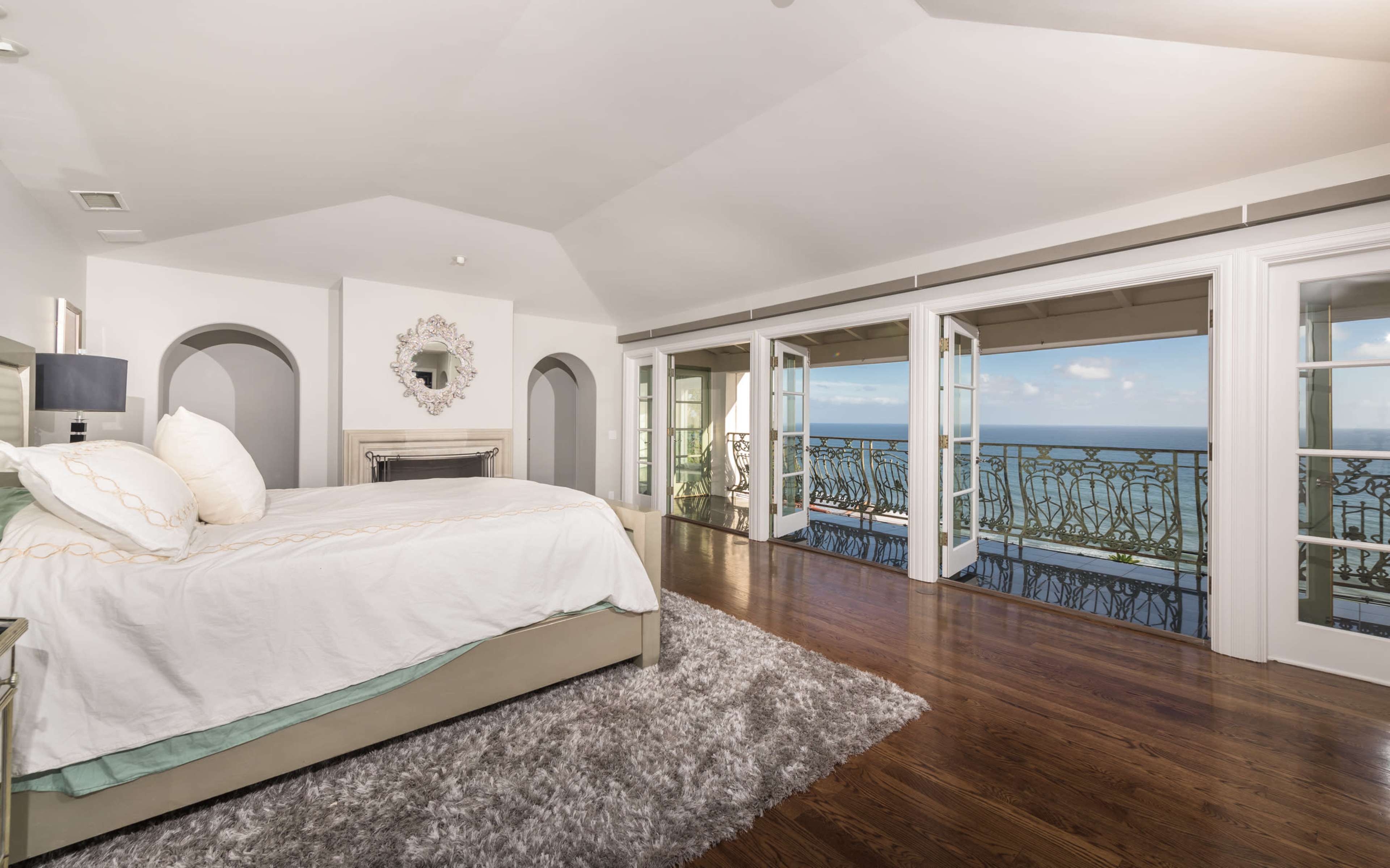 The image shows a spacious bedroom with a large bed and a balcony that offers a view of the ocean.