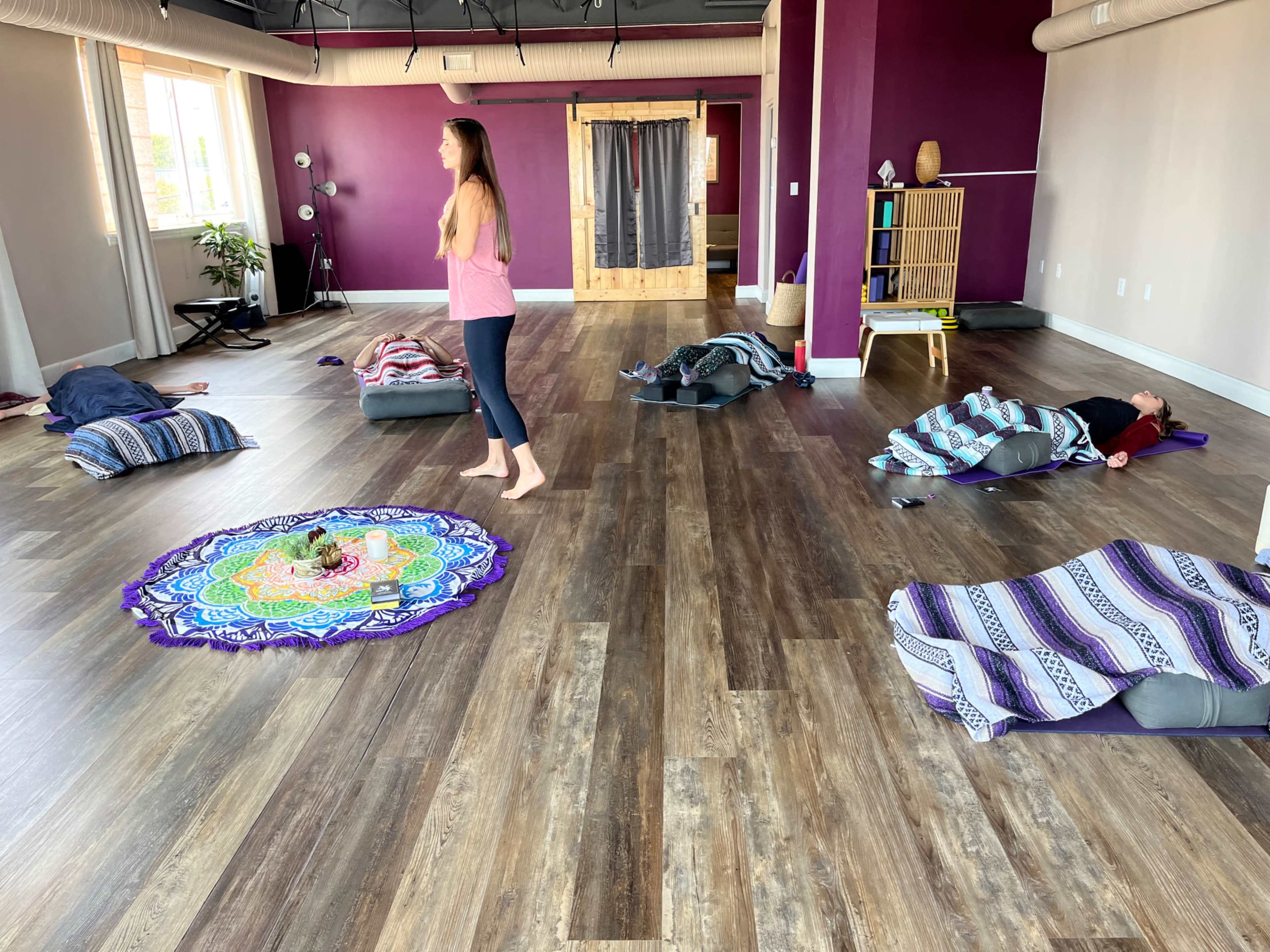 A yoga studio features individuals lying on mats covered with blankets while a person stands in front, leading a session.
