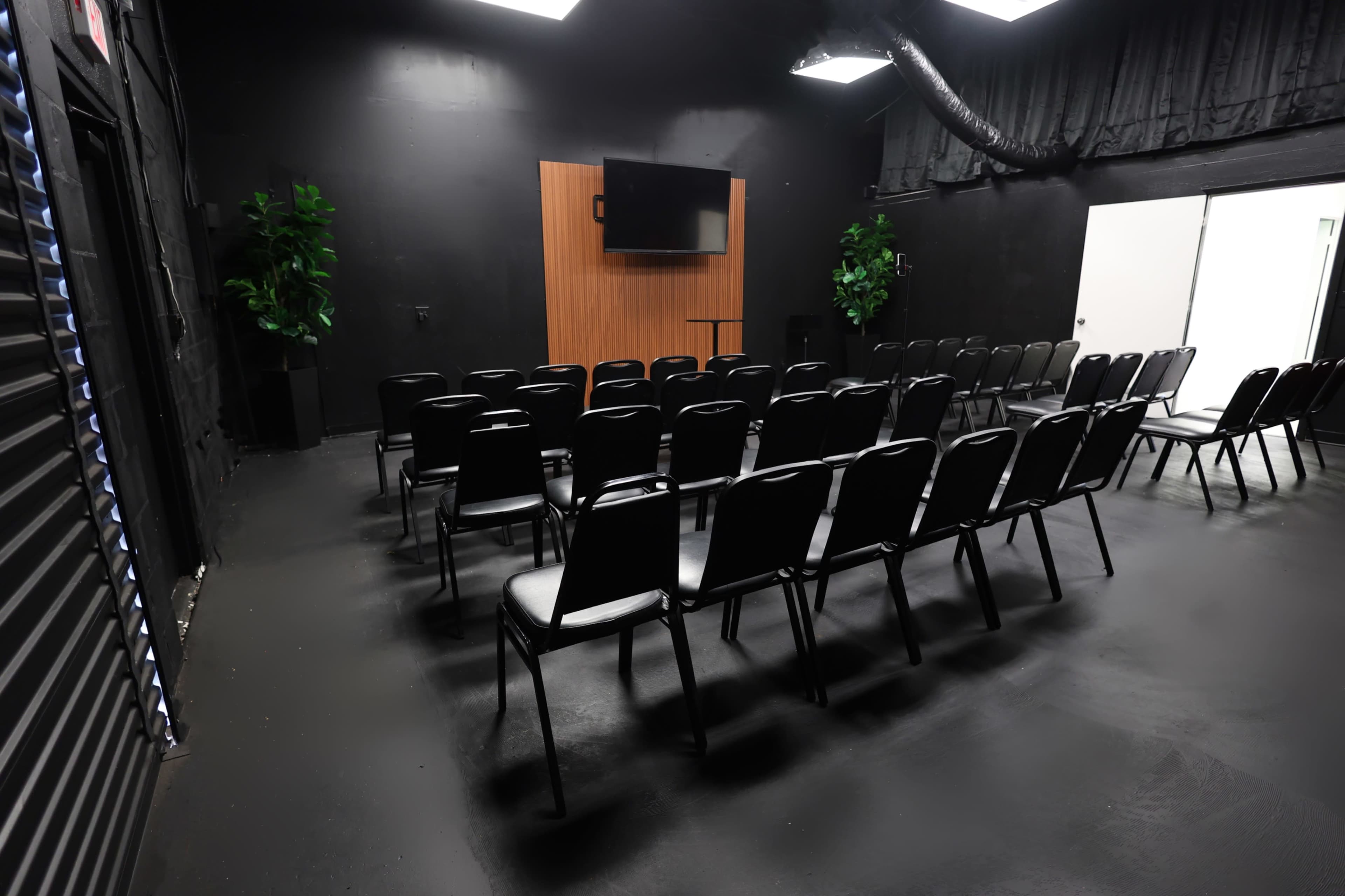 The image shows a black-walled meeting room set up with rows of black chairs facing a wooden panel with a television mounted on it.