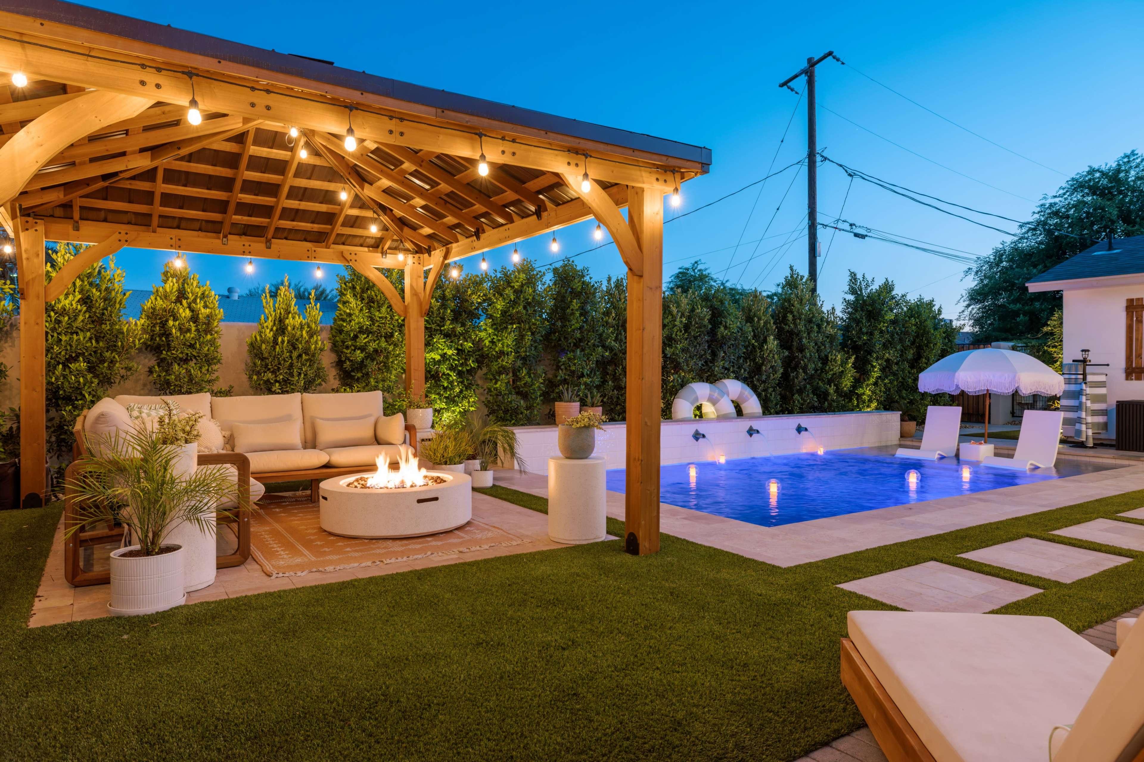 The image shows a landscaped backyard featuring a wooden pergola with seating and a fire pit, alongside a pool and lounge chairs under evening lighting.