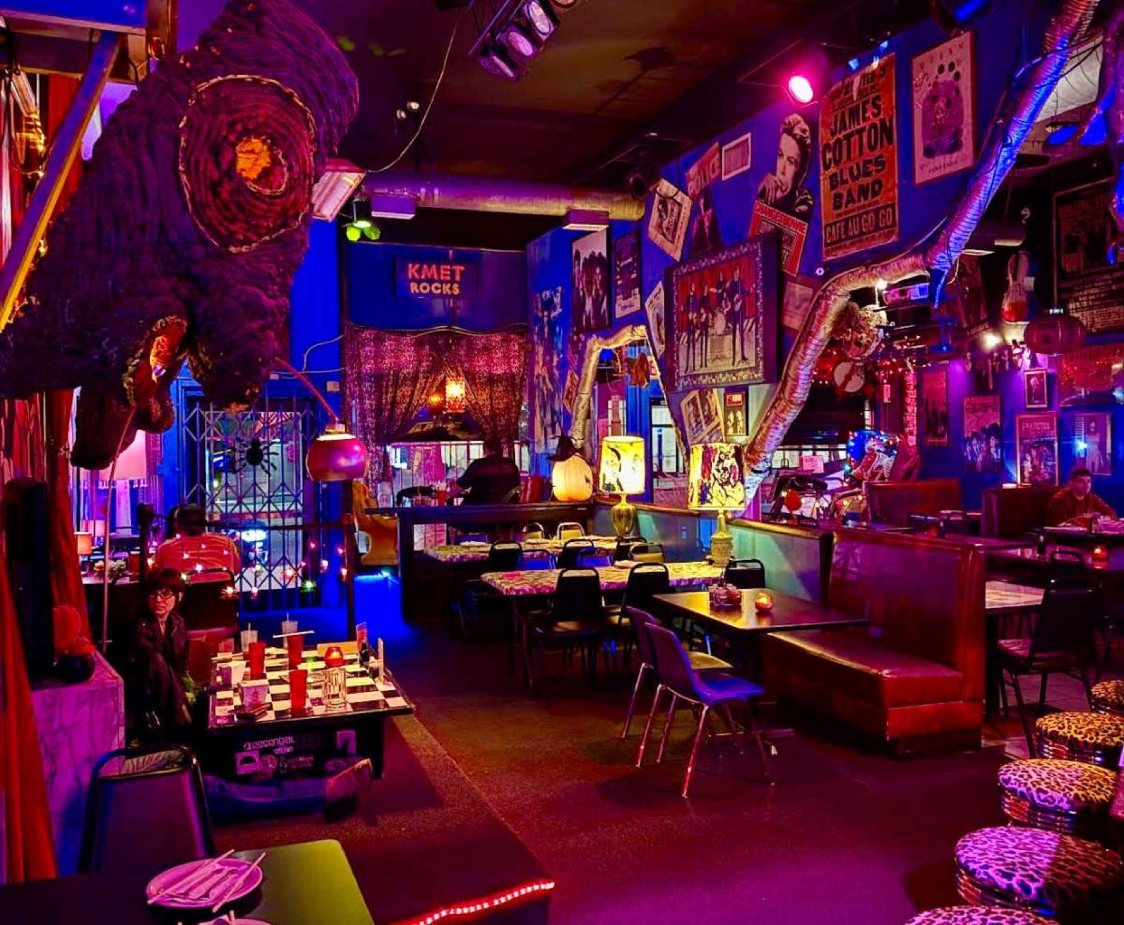 The image shows a vibrant bar interior adorned with colorful lighting, eclectic decorations, and a mix of seating arrangements, including booths and tables.