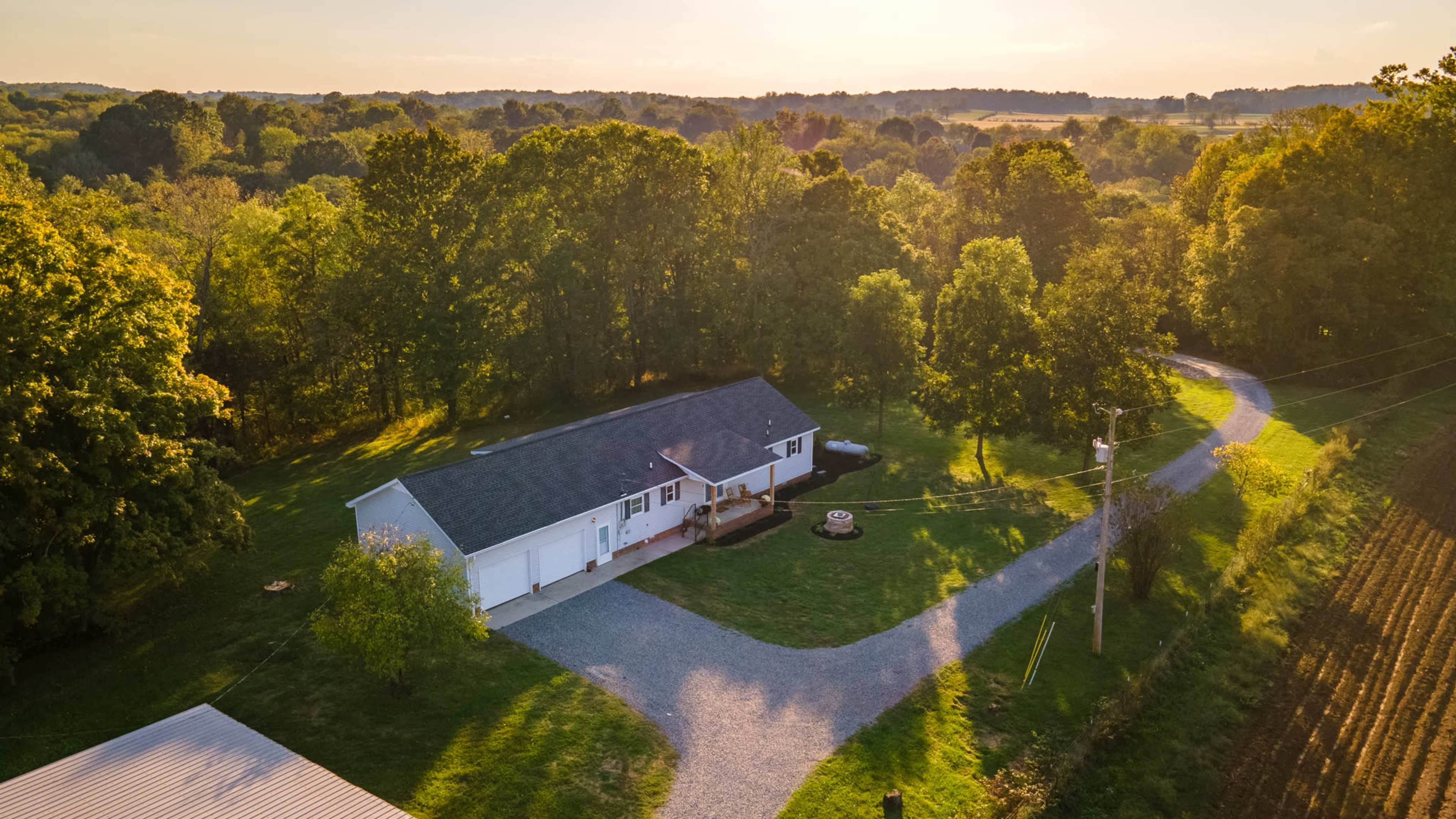 A single-story house with a gravel driveway is surrounded by trees and fields in a rural setting during sunset.