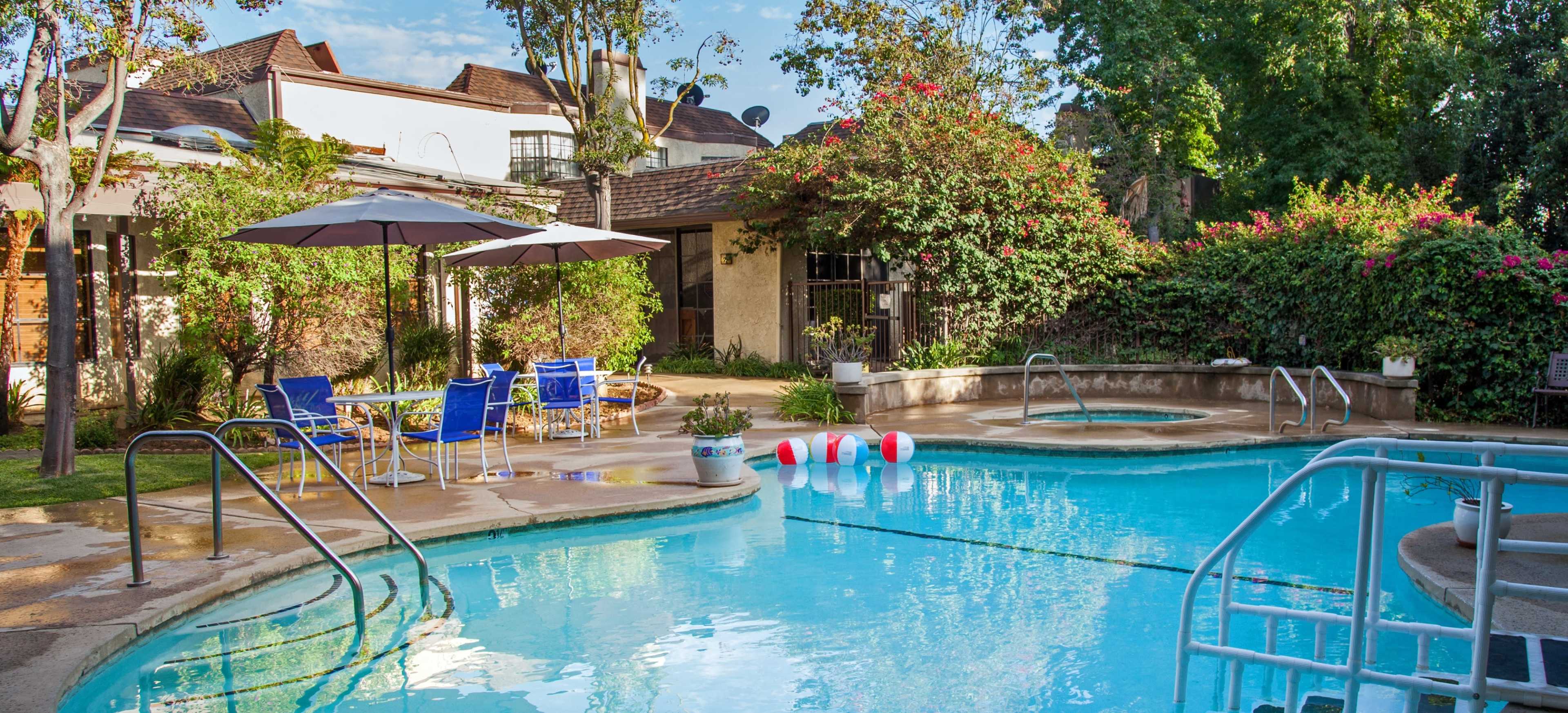 The image shows a pool area with blue water, several beach balls floating, surrounded by greenery, lounge chairs, and an umbrella.