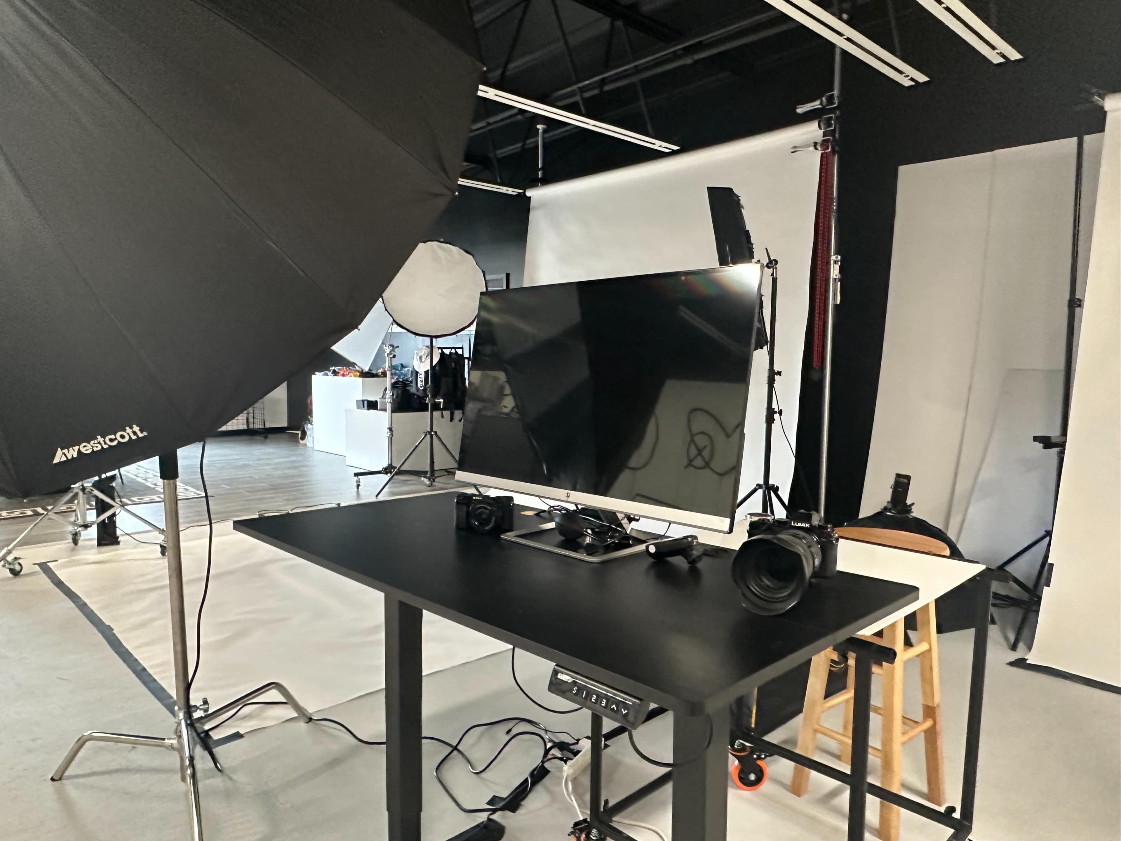 A photography studio setup features a large monitor and cameras on a table, with lighting equipment and backdrops in the background.