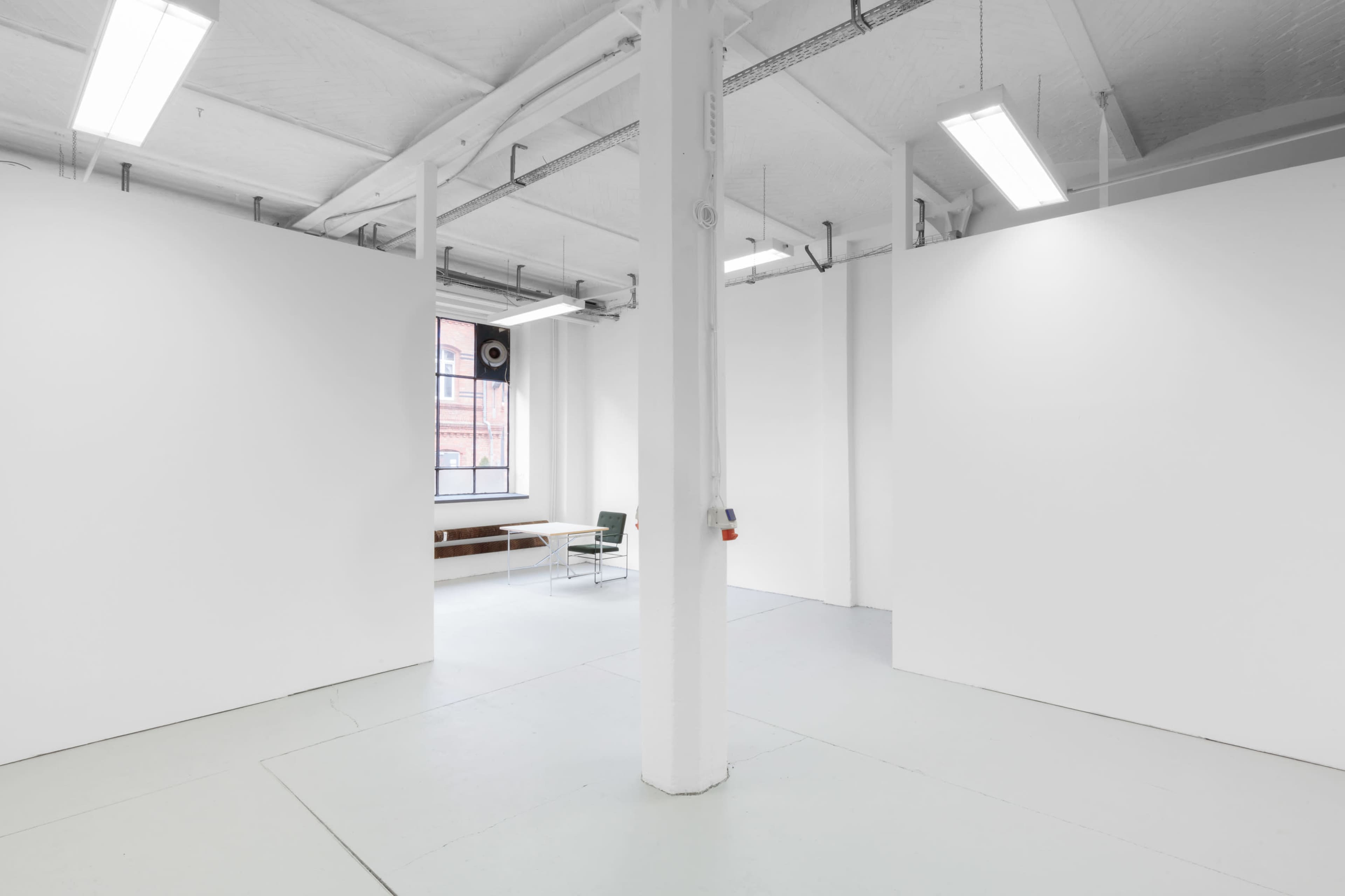 A spacious, empty room with white walls, a single central column, and a chair and table near a window.