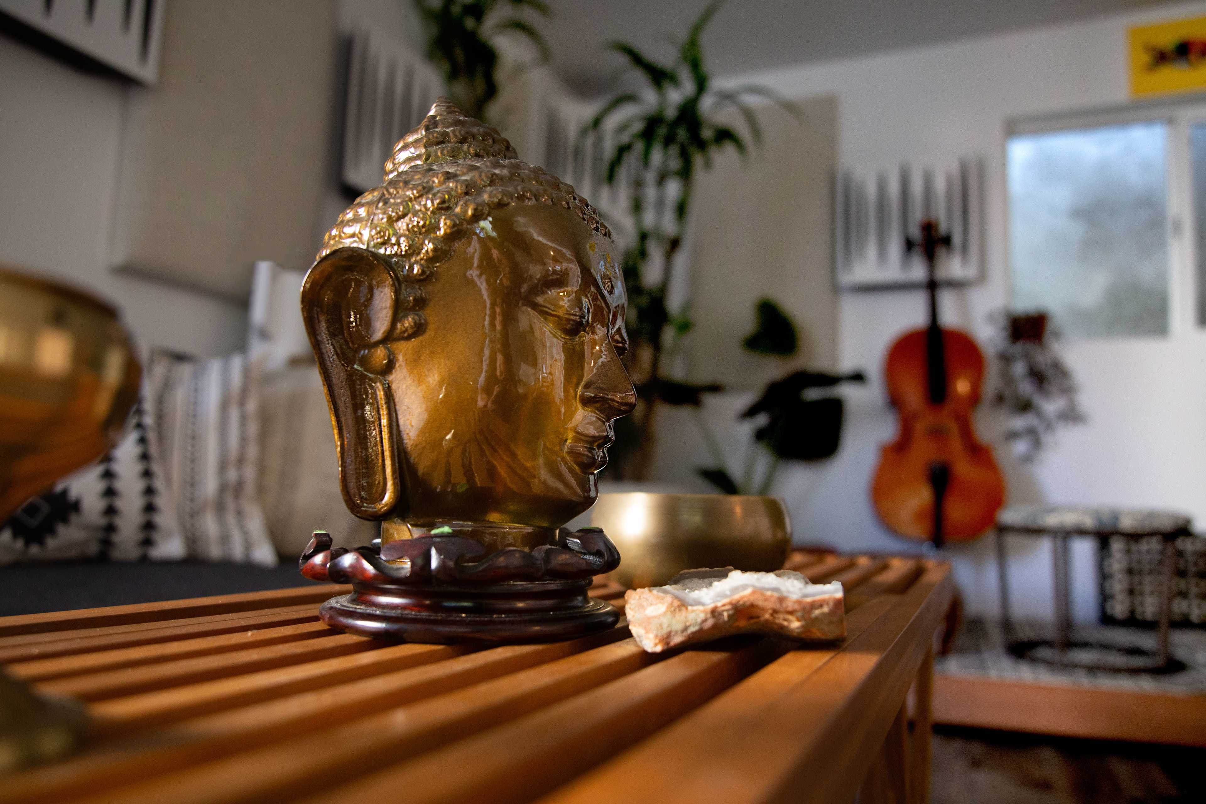 A bronze Buddha head sculpture sits on a wooden table beside a small stone and a bowl in a room decorated with plants and musical instruments.