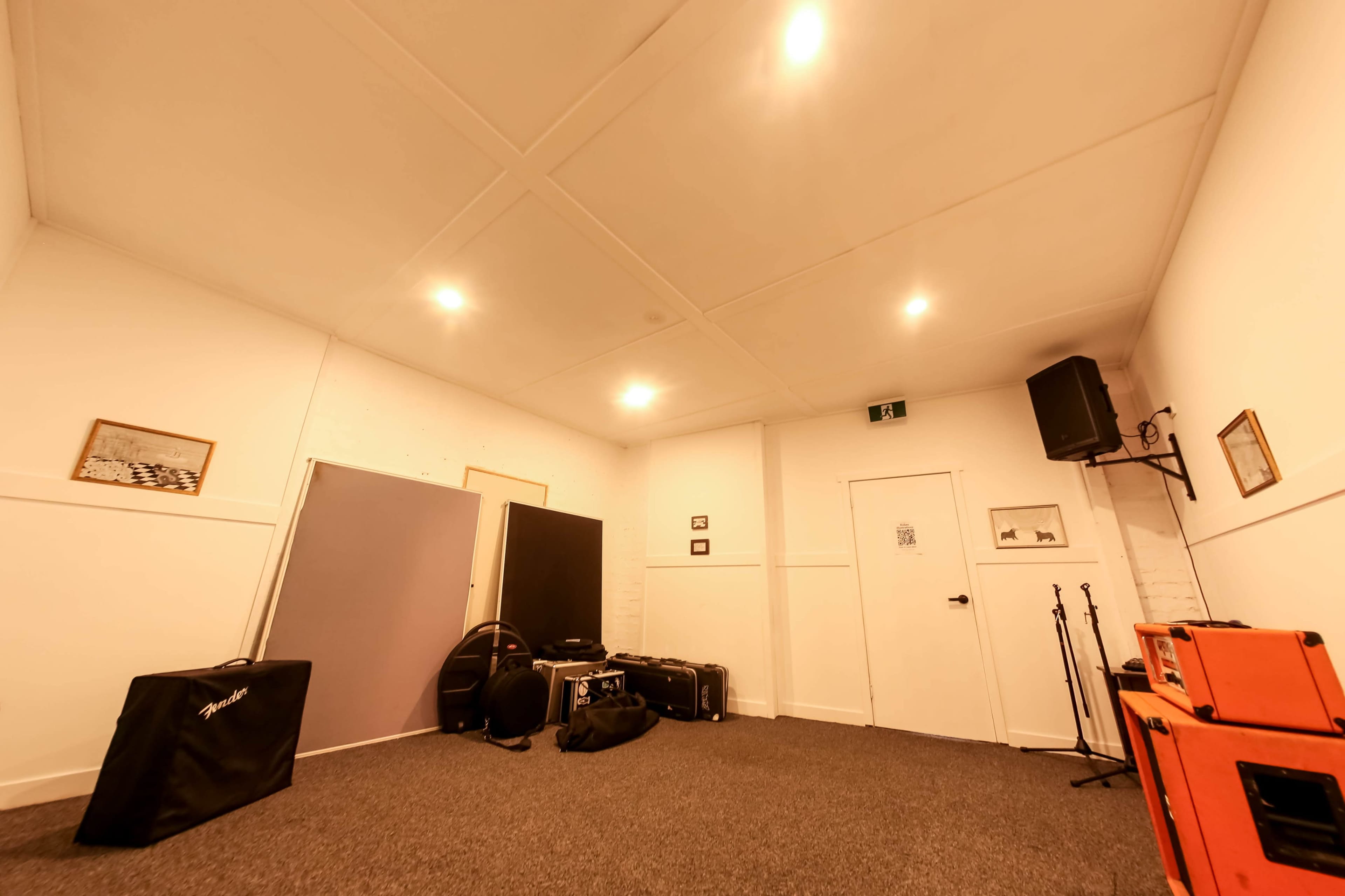The image shows a soundproof rehearsal room featuring a carpeted floor, various equipment cases, and wall-mounted speakers.