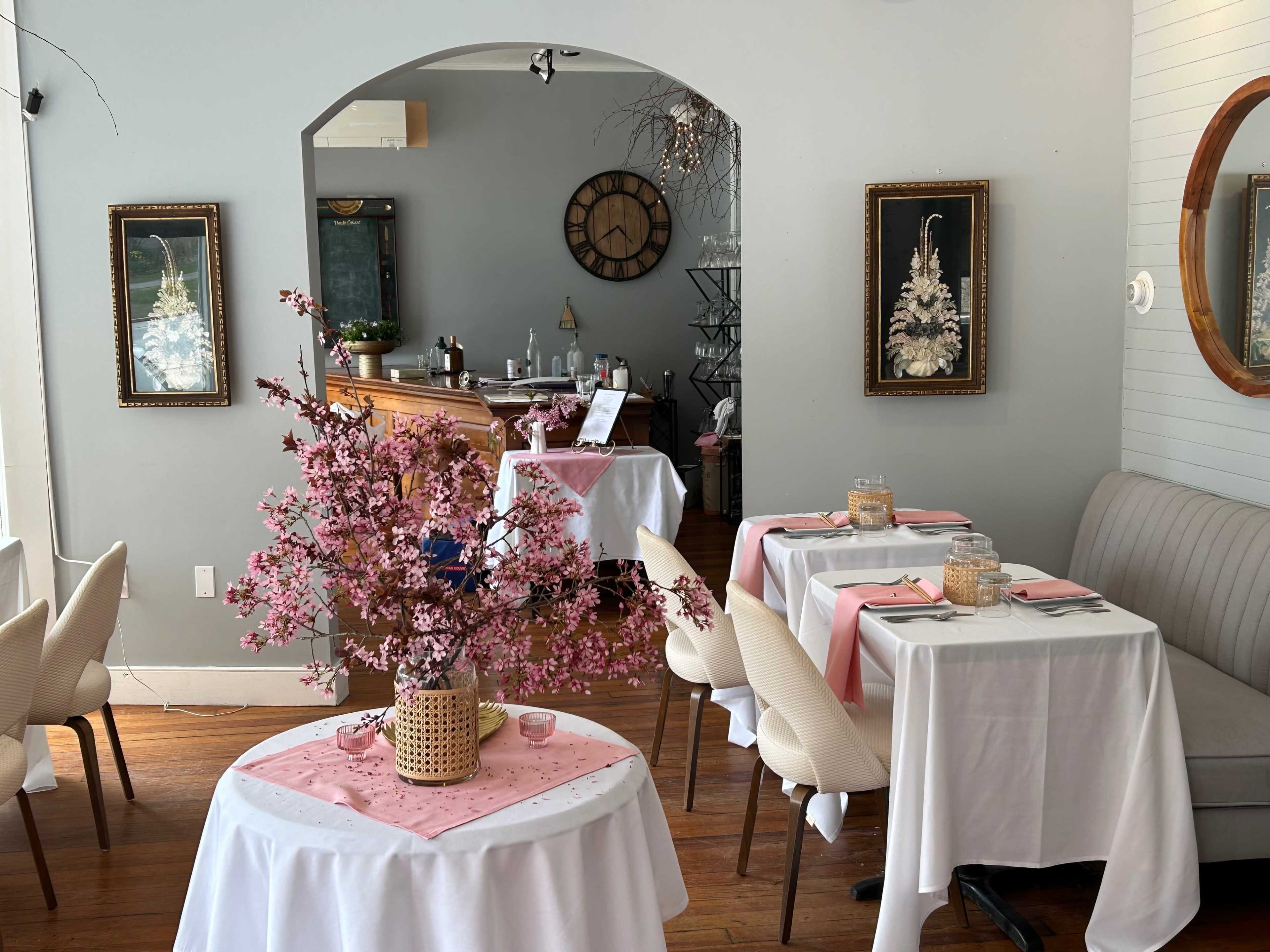 A cozy restaurant interior with white tablecloths, pink accents, and a decorative cherry blossom centerpiece.