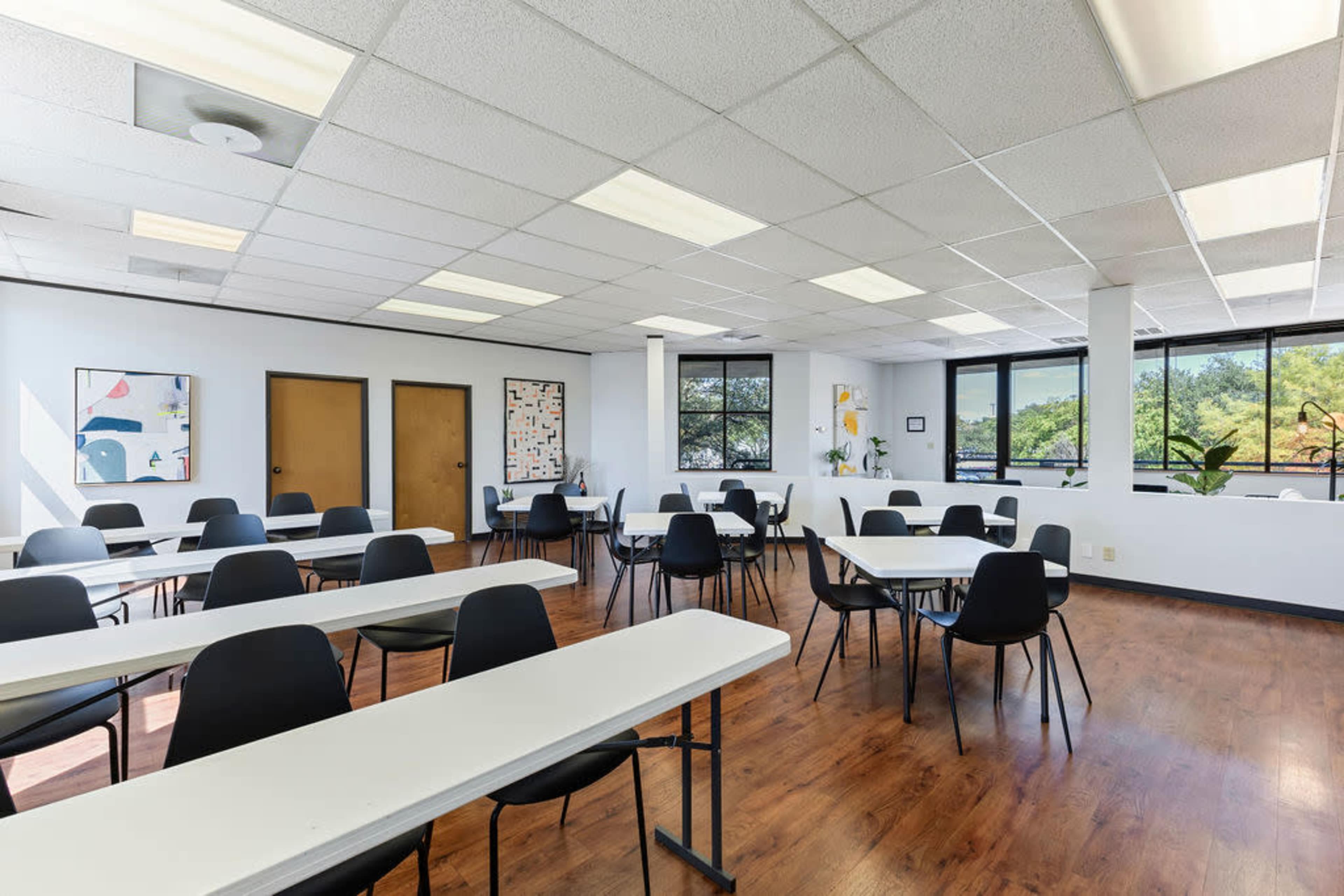A spacious classroom features multiple tables and chairs arranged for a group setting, with windows allowing natural light to enter.