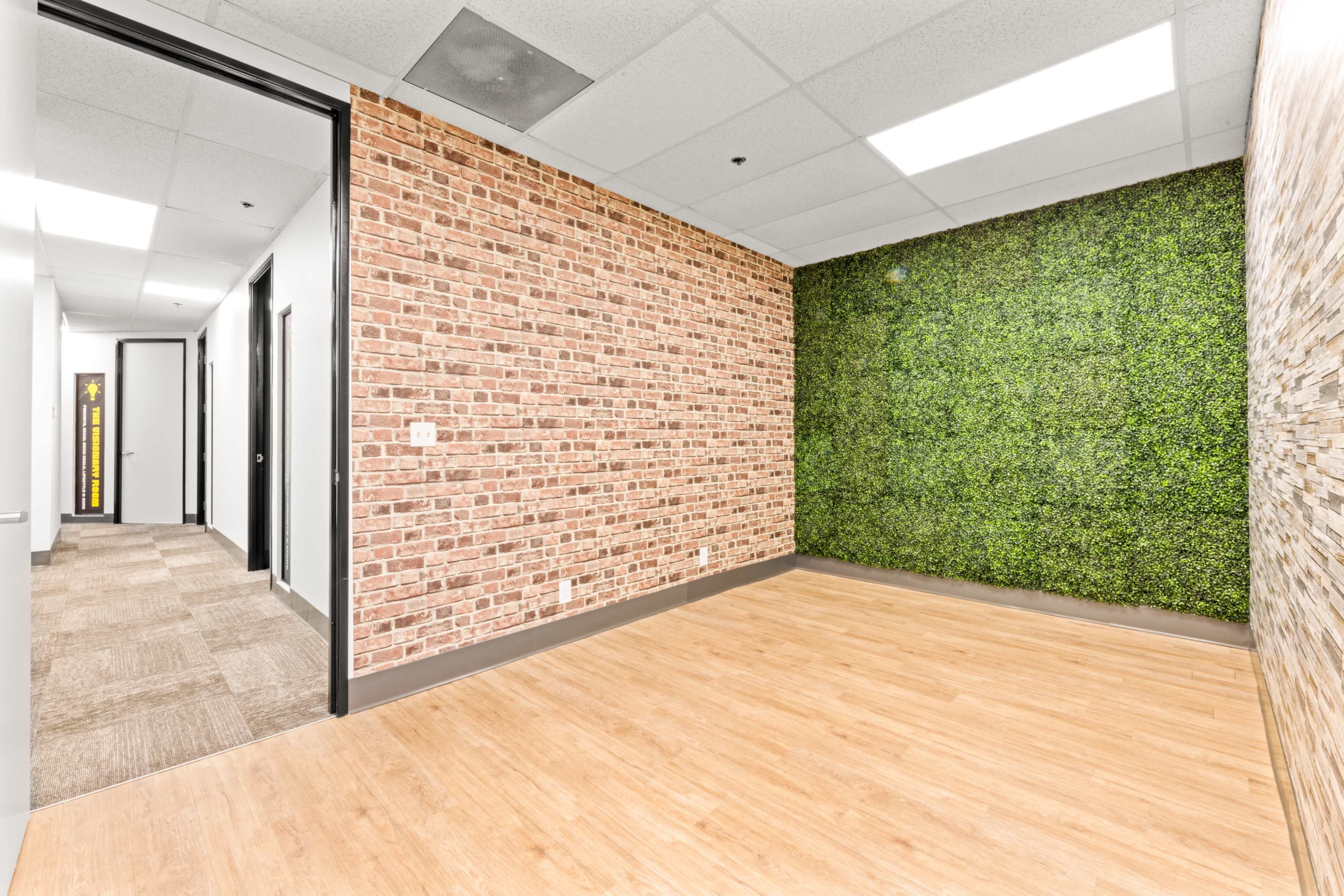The image shows a vacant room with one wall featuring a brick pattern and another wall covered in green plant-like decor.