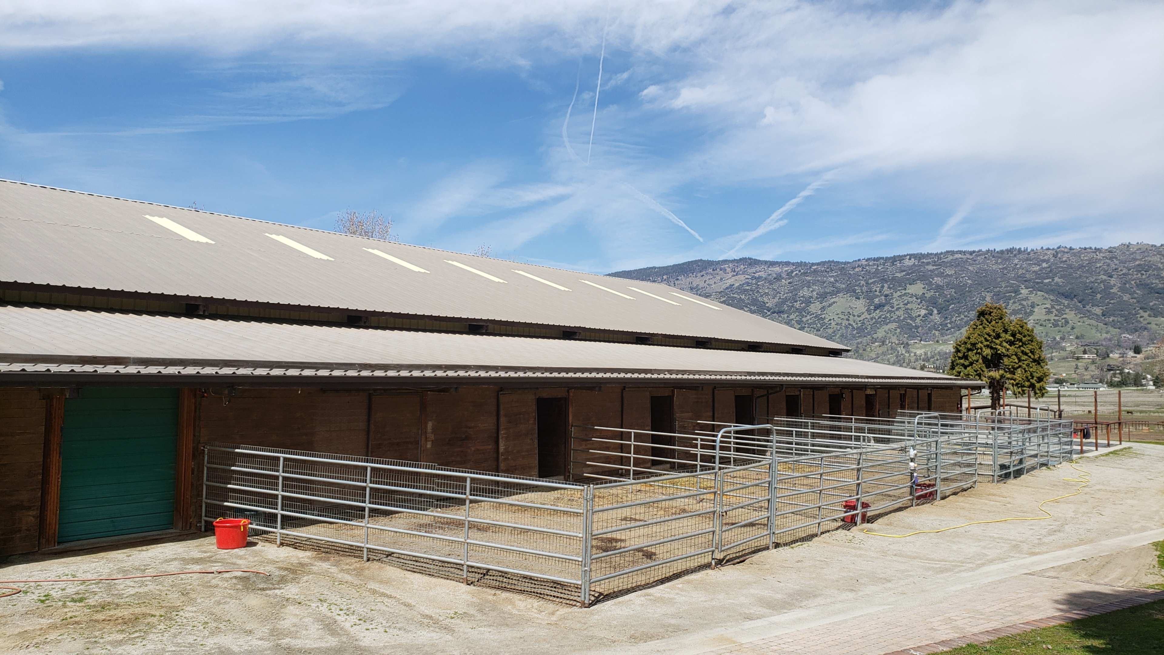 A barn with multiple horse stalls and empty paddocks against a mountainous background under a blue sky.