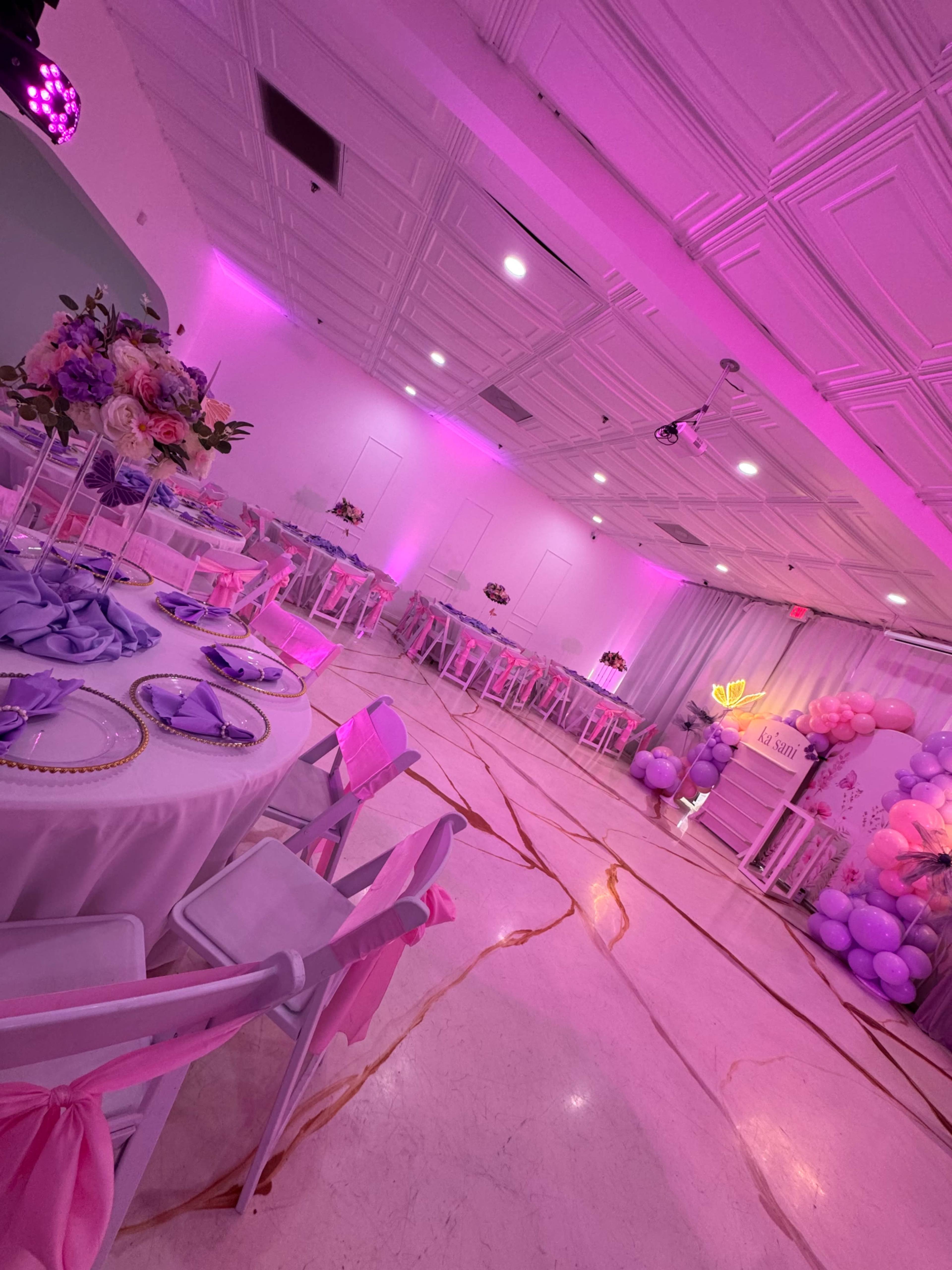The image depicts a decorated event space featuring pink lighting, elegant tables set with plates and napkins, and balloon arrangements in one corner.