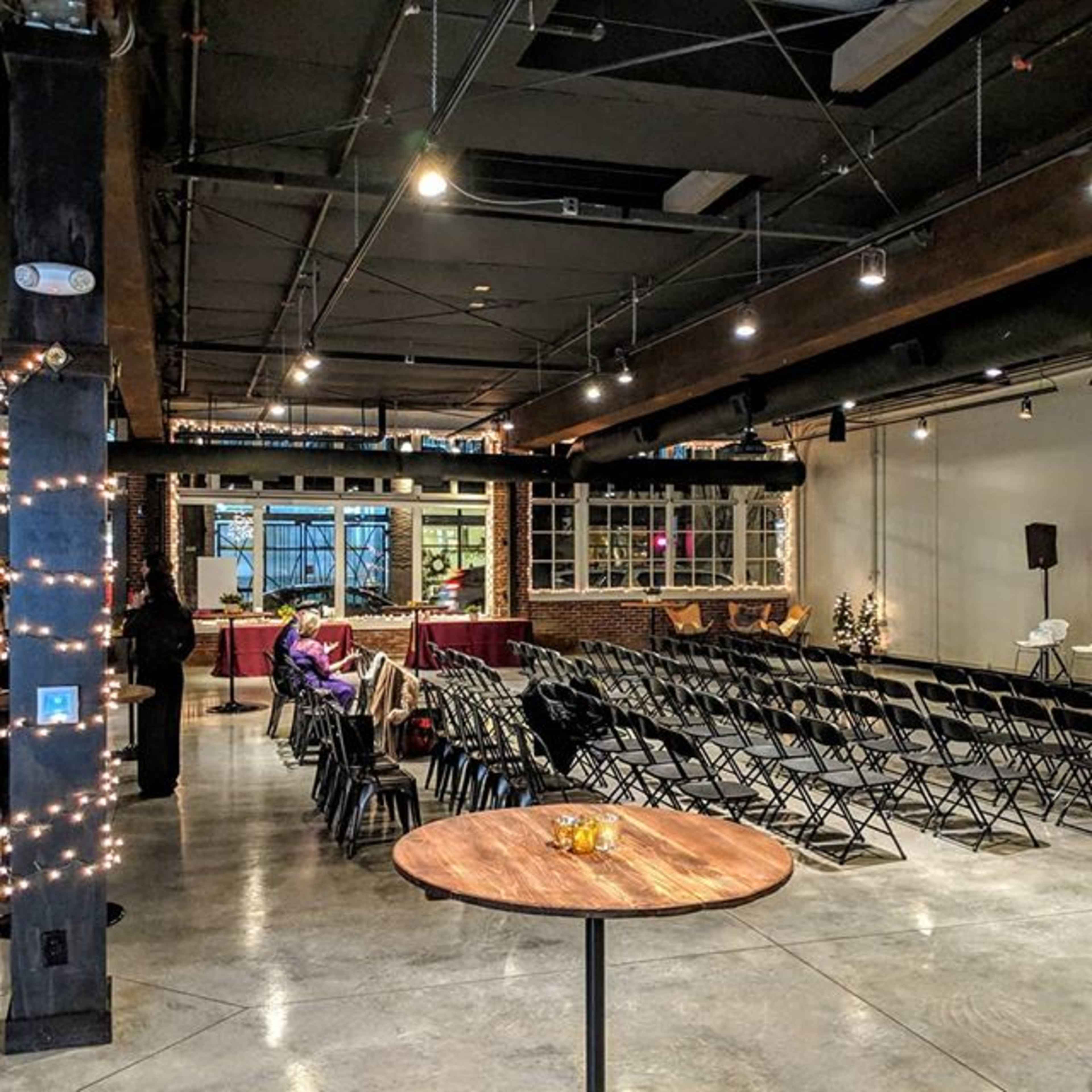 The image shows a spacious event venue with rows of black folding chairs arranged facing a small stage and a wooden table in the foreground.