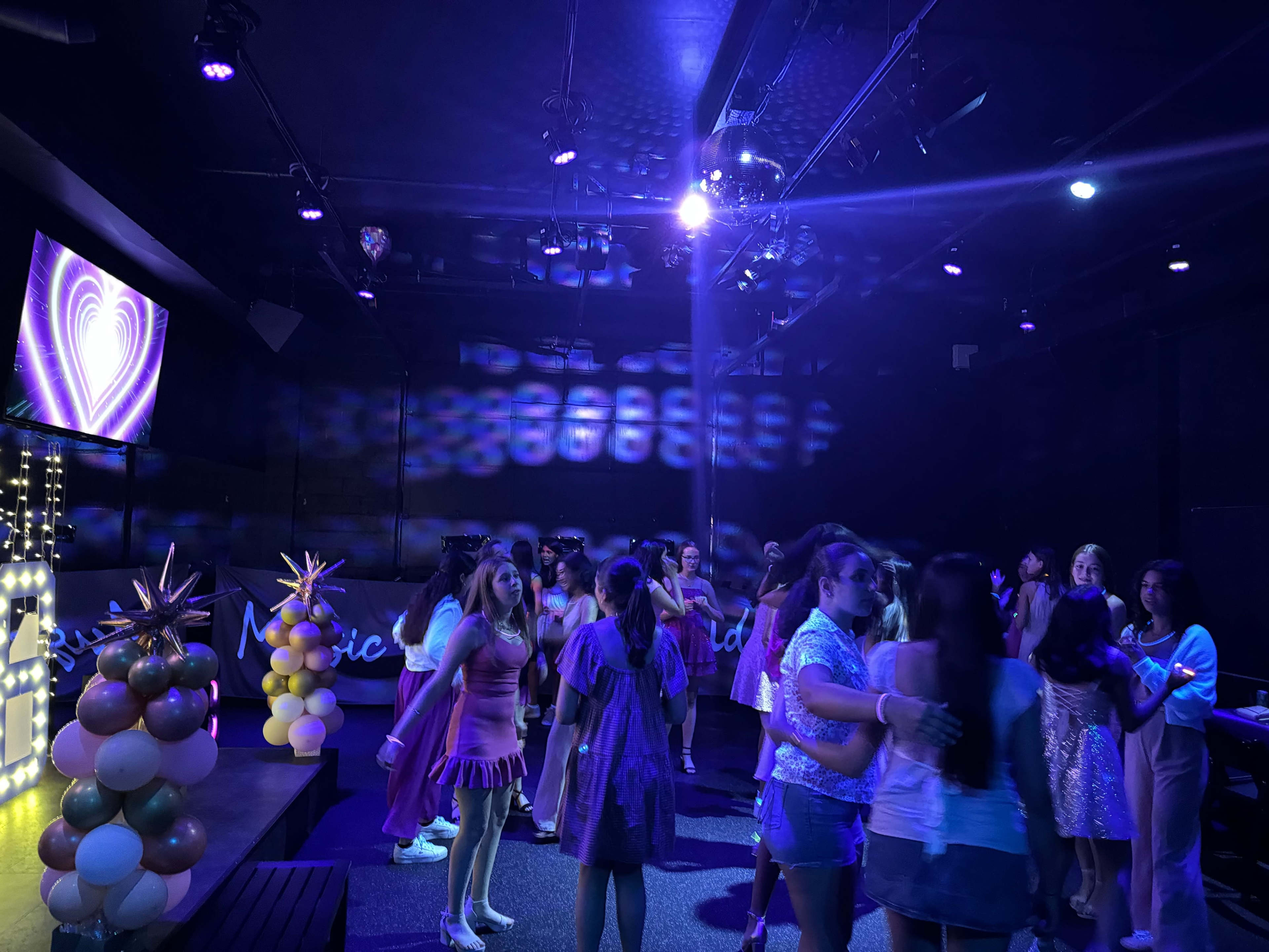 A group of young people dance and socialize in a dimly lit venue decorated with colorful lights and balloons.