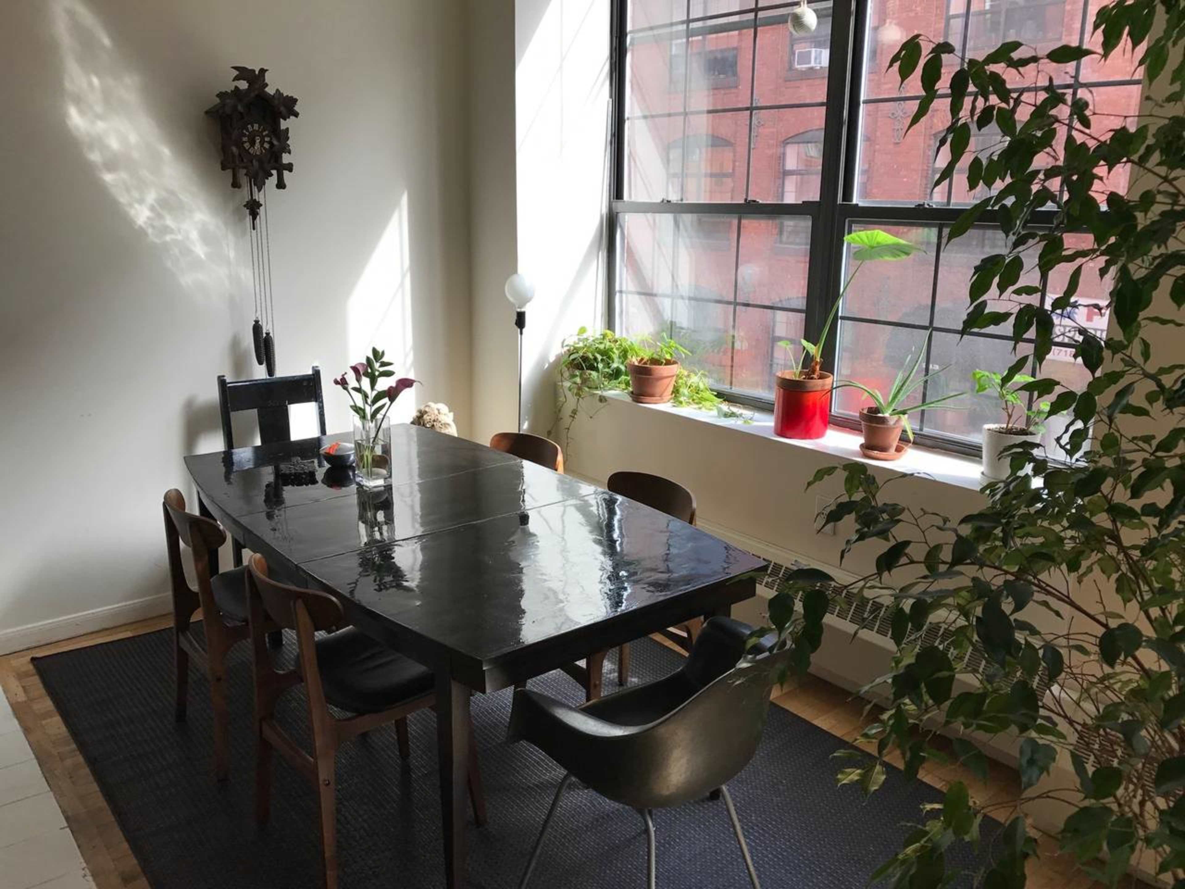 A rectangular dining table with six chairs is arranged in a bright room featuring large windows and potted plants.