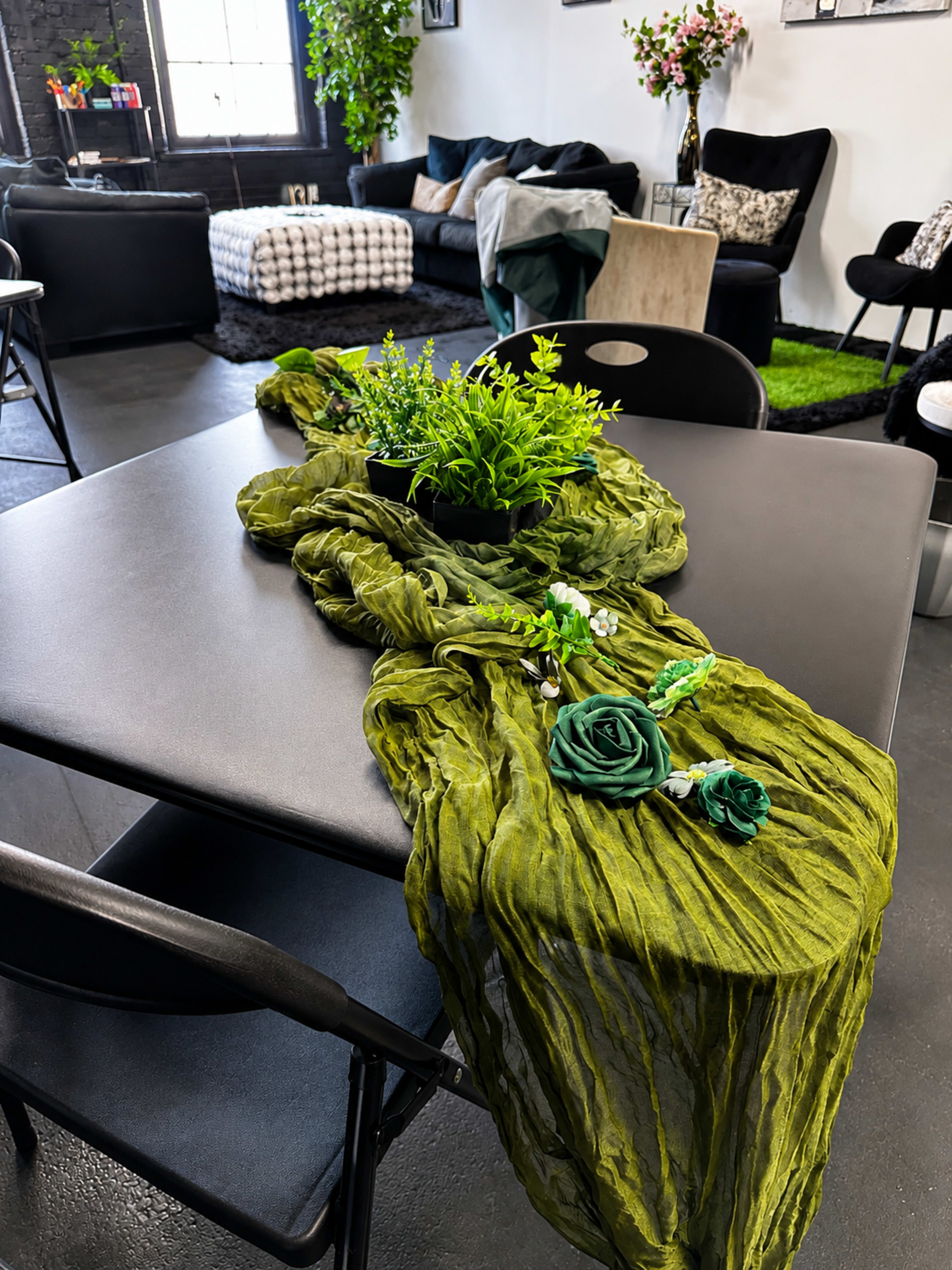 A long, green fabric drapes across a black table, accompanied by a small planter with greenery and decorative floral accessories, set in a stylish room with modern furniture.