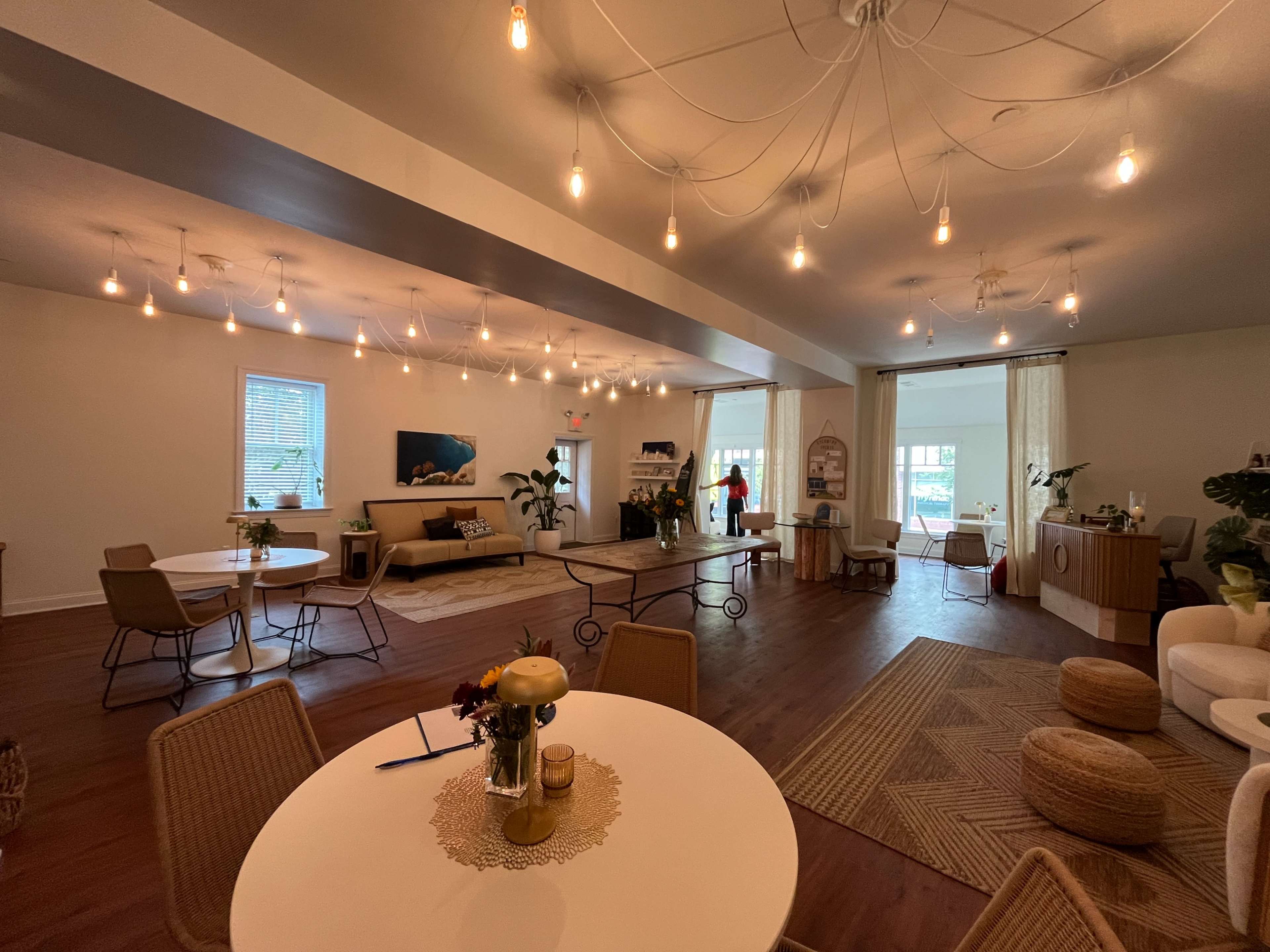 The image shows a spacious, well-lit interior with several tables and chairs, decorative plants, and a cozy seating area featuring a sofa.